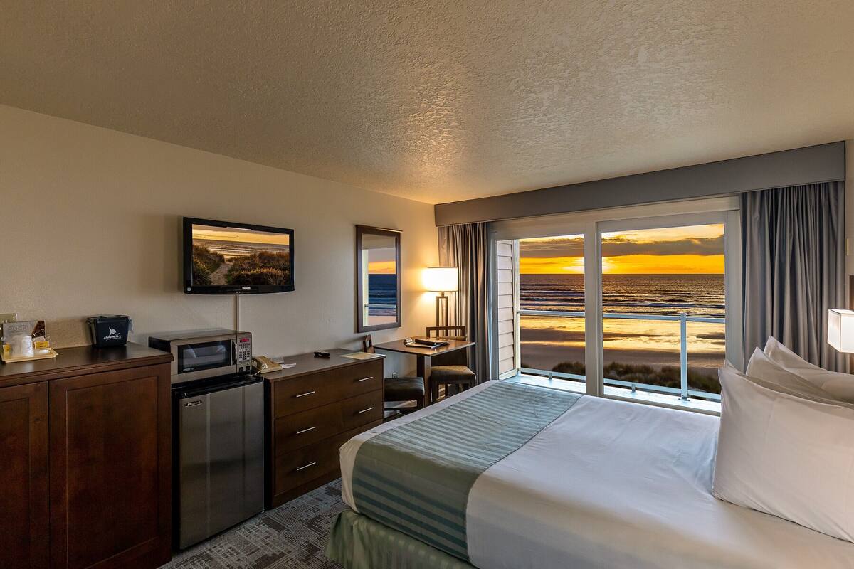 A hotel room with a bed, a desk, a refrigerator, a microwave, a television, and a large window with a view of the beach at sunset.