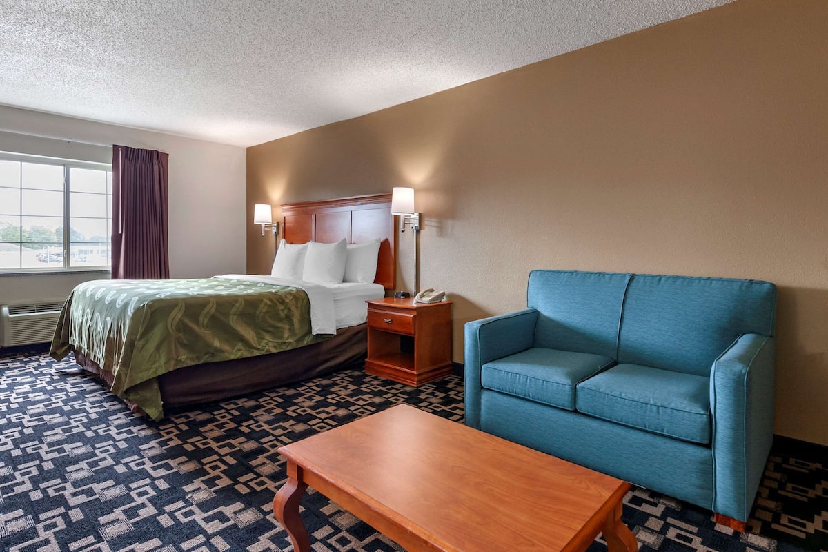 Una habitación de hotel con una cama, un sillón azul, una mesita de noche de madera con una lámpara y una alfombra estampada.