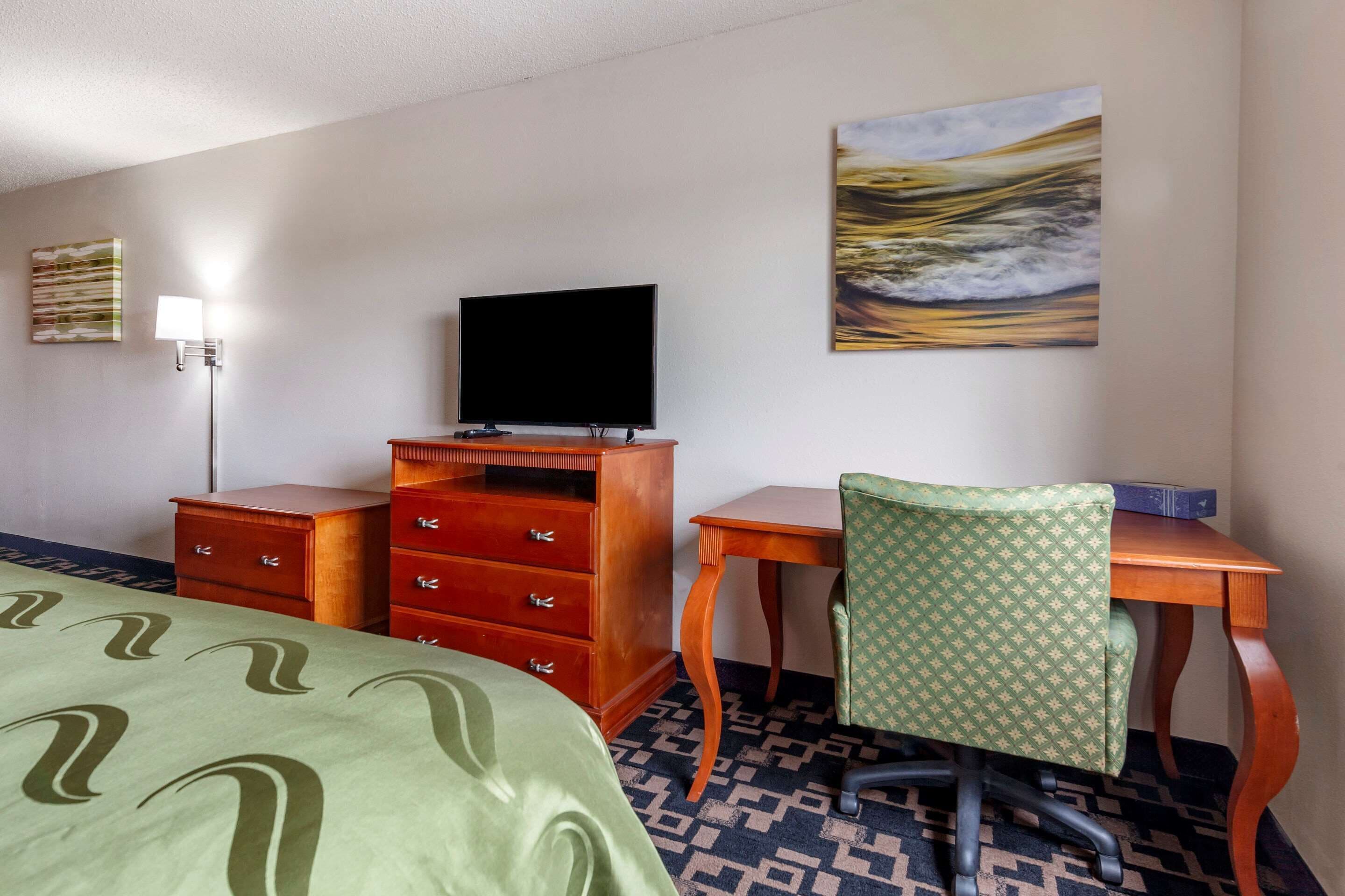 A hotel room with a bed, a desk with a chair, a dresser, a flat-screen TV, and a painting on the wall.