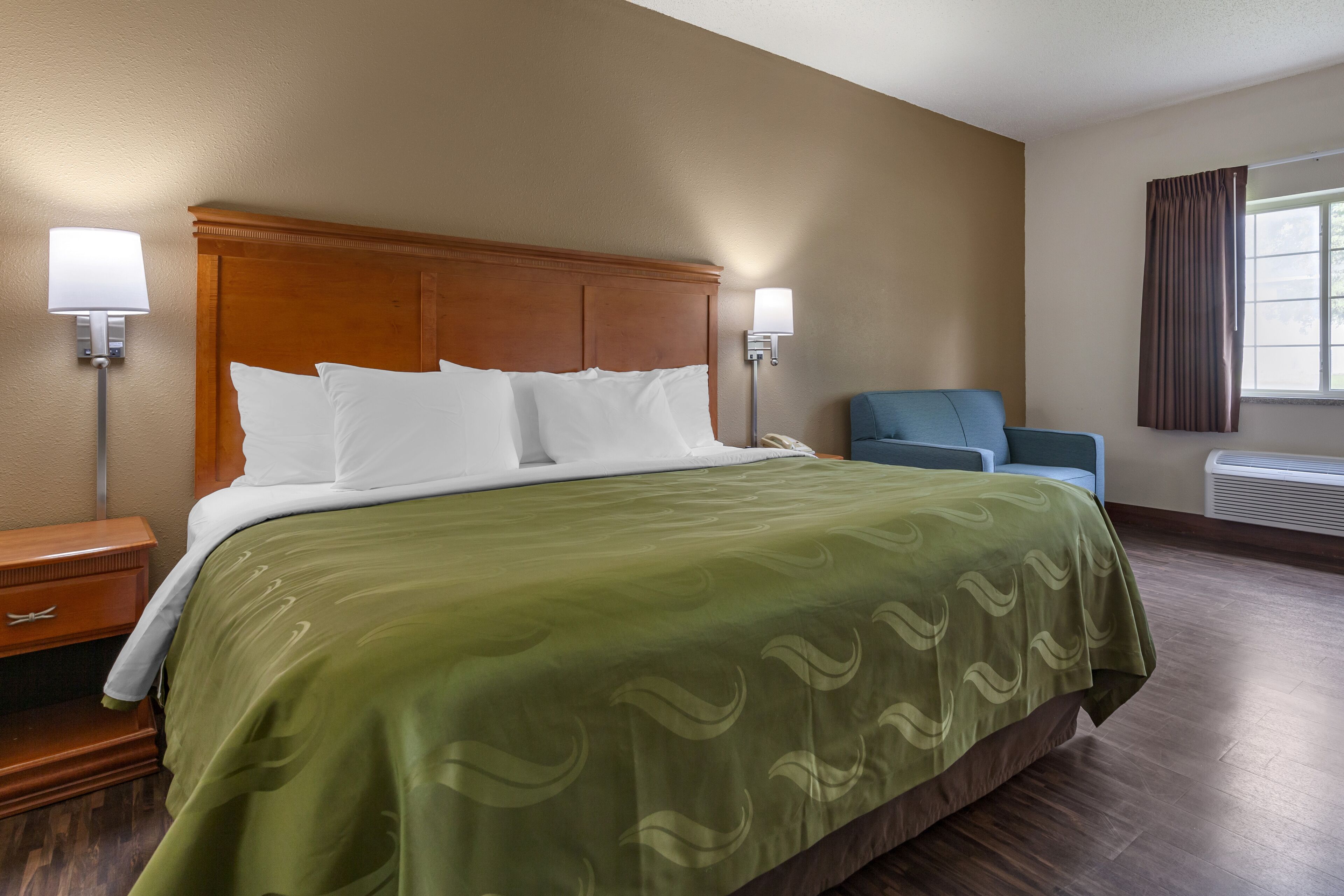 A hotel room with a large bed, two bedside lamps, a blue armchair, and a window with curtains.