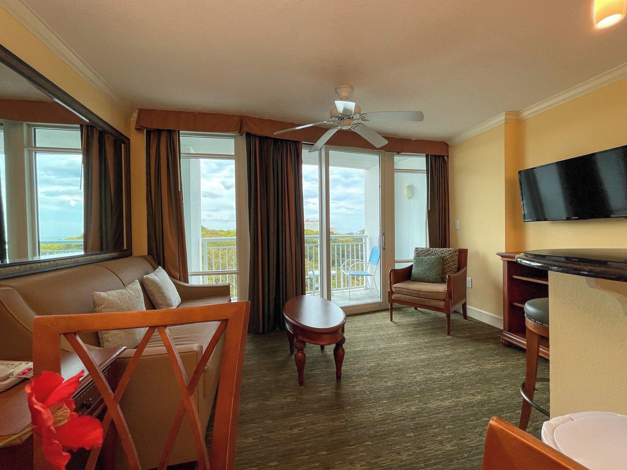 A hotel room with a sofa, a small table, a chair, a ceiling fan, a TV, and a balcony with a view.