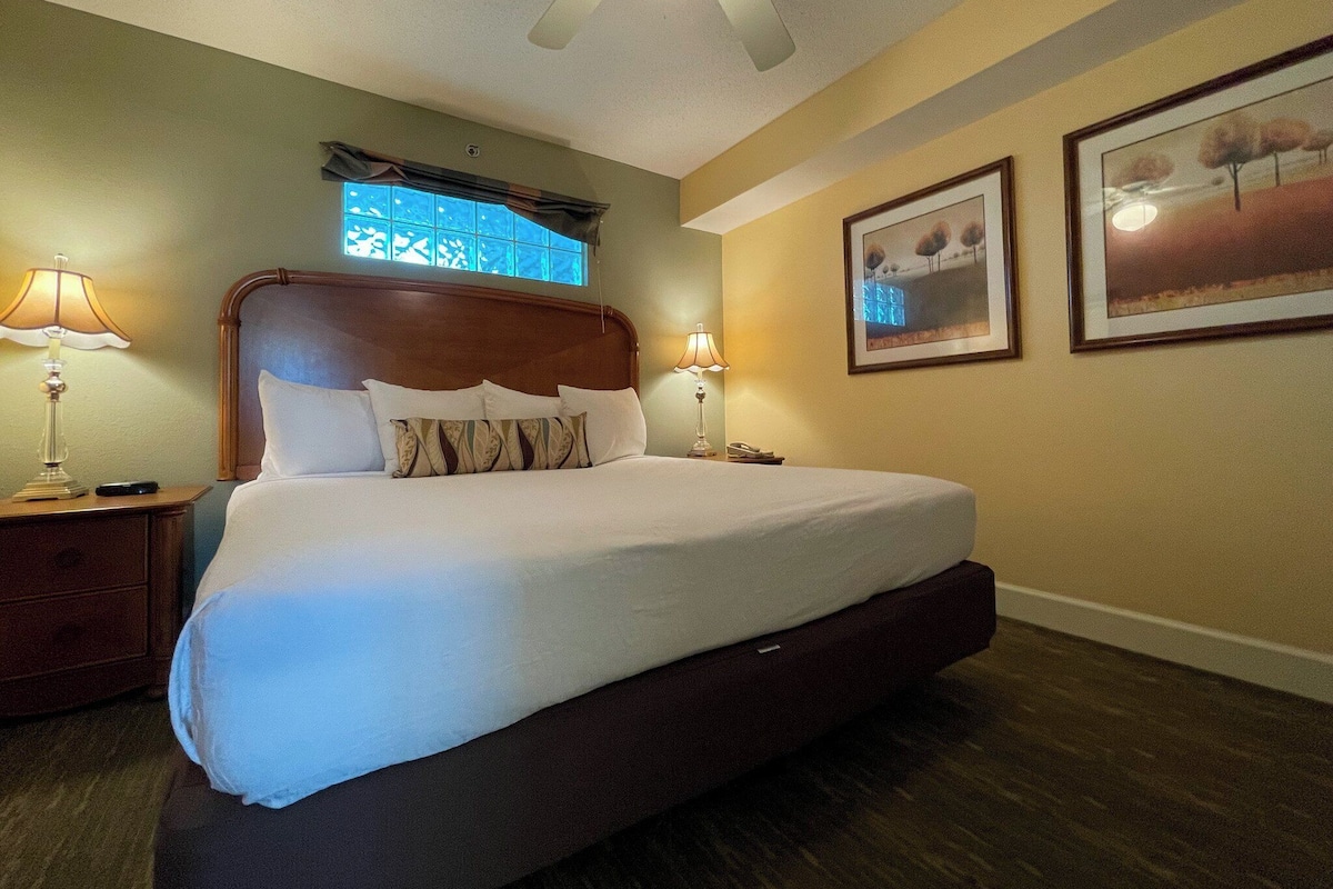 Quarto de hotel com uma cama grande, duas mesas de cabeceira, um ventilador de teto e duas imagens emolduradas na parede.