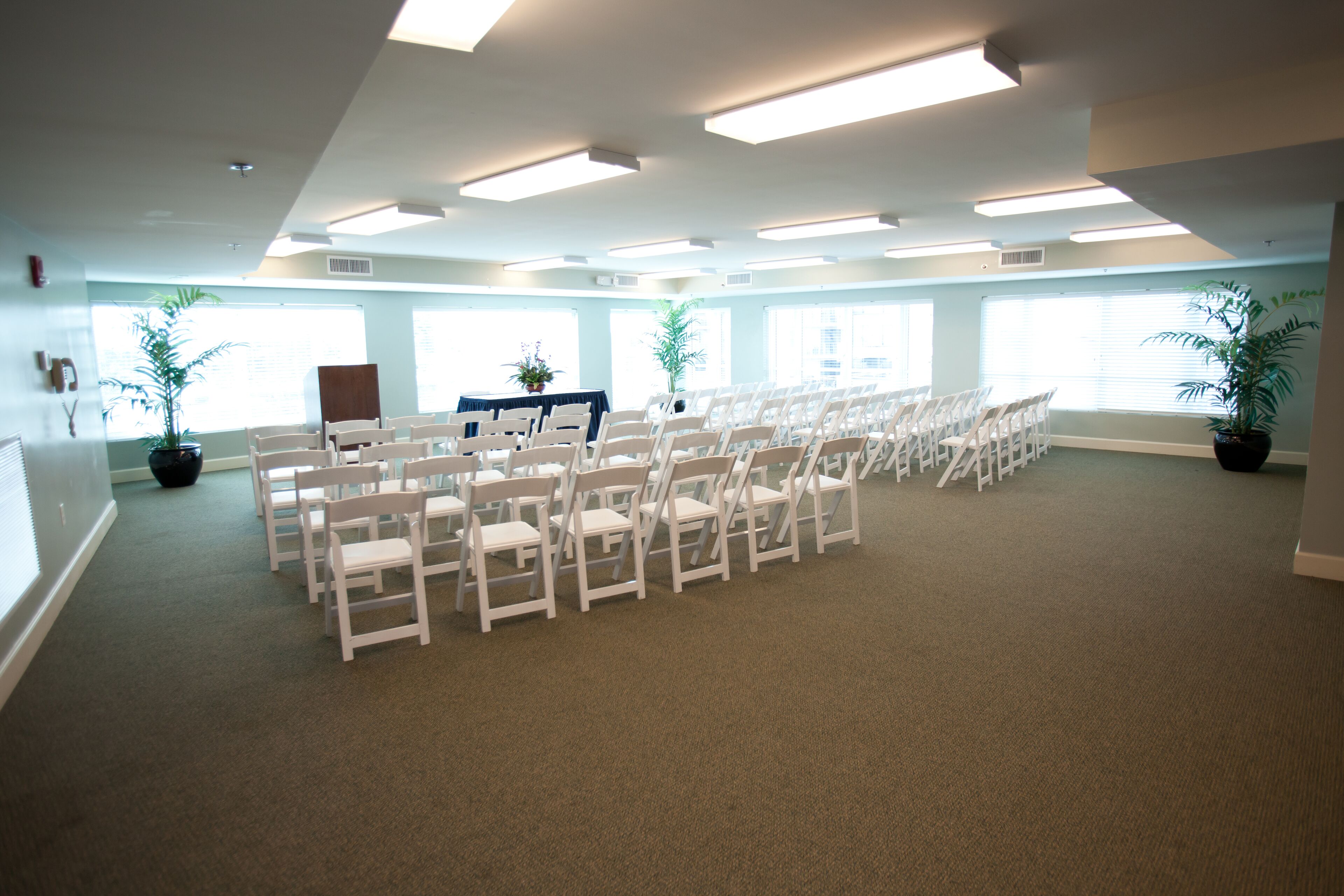 A conference room with rows of white chairs, large windows, and ceiling lights.