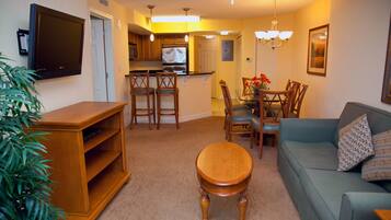 A living room with a sofa, a coffee table, and a TV mounted on the wall. There is a dining area with a table and chairs, and a kitchen in the background.