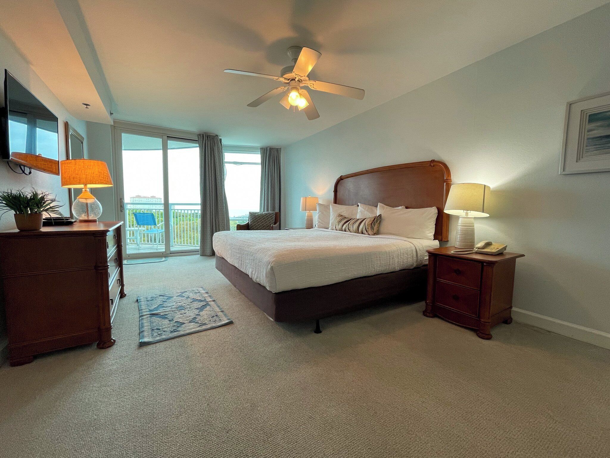 Une chambre spacieuse avec un grand lit, un ventilateur de plafond, un bureau avec une lampe et une vue sur l’extérieur grâce à la porte-fenêtre coulissante.