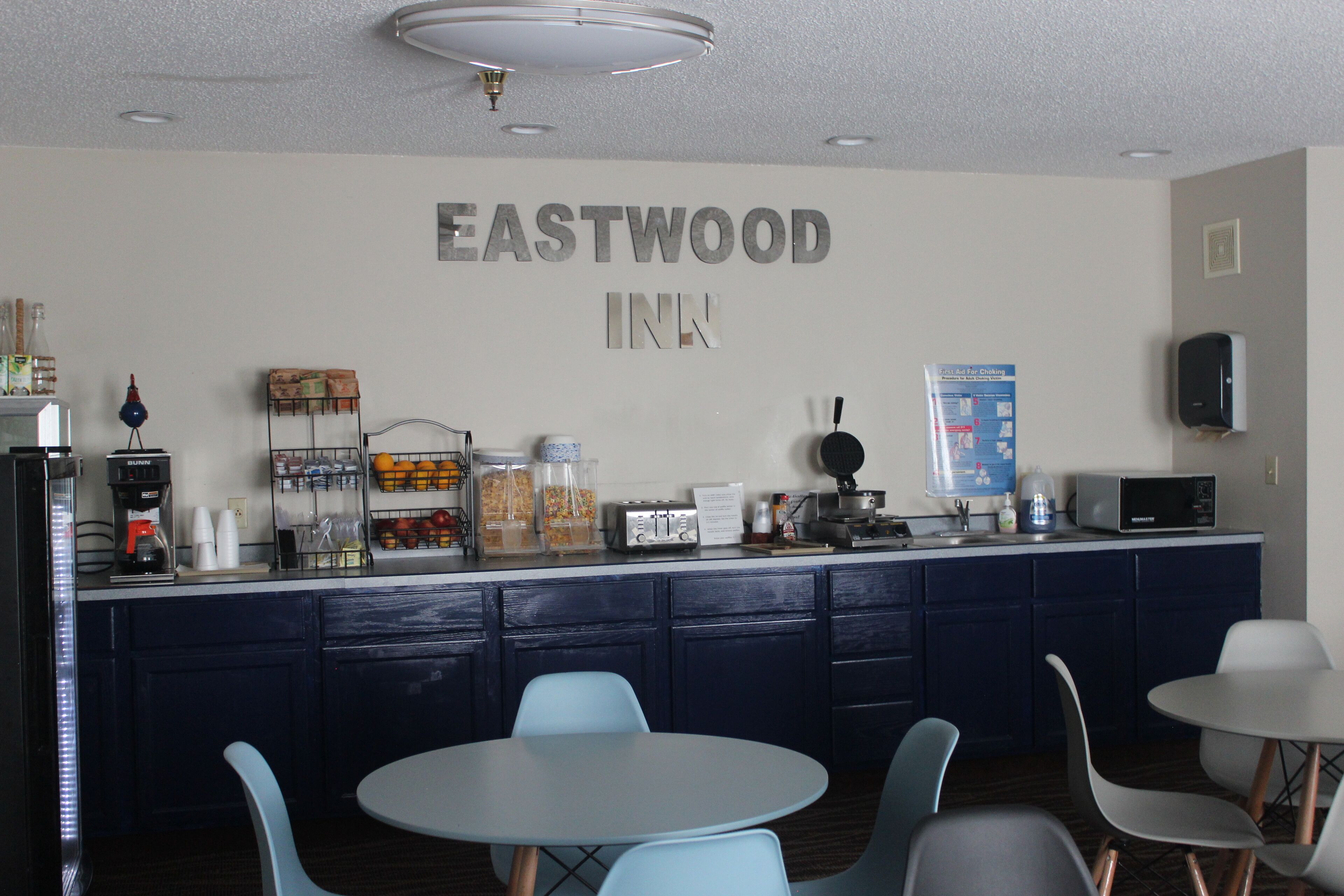 Un área de kitchenette con cafetera, tostadora y microondas. Hay sillas azules y una pequeña mesa en primer plano. La pared tiene un letrero que dice "EASTWOOD INN".