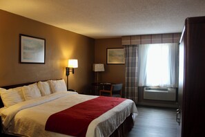A hotel room with a bed, a desk, a chair, a window with curtains, and an air conditioning unit.