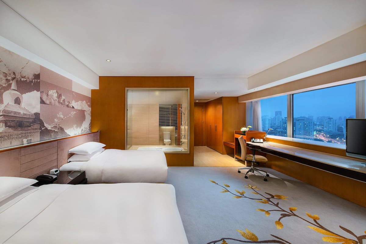 Habitación de hotel con dos camas, un escritorio, un ventanal grande con vistas a la ciudad y un mural en la pared.
