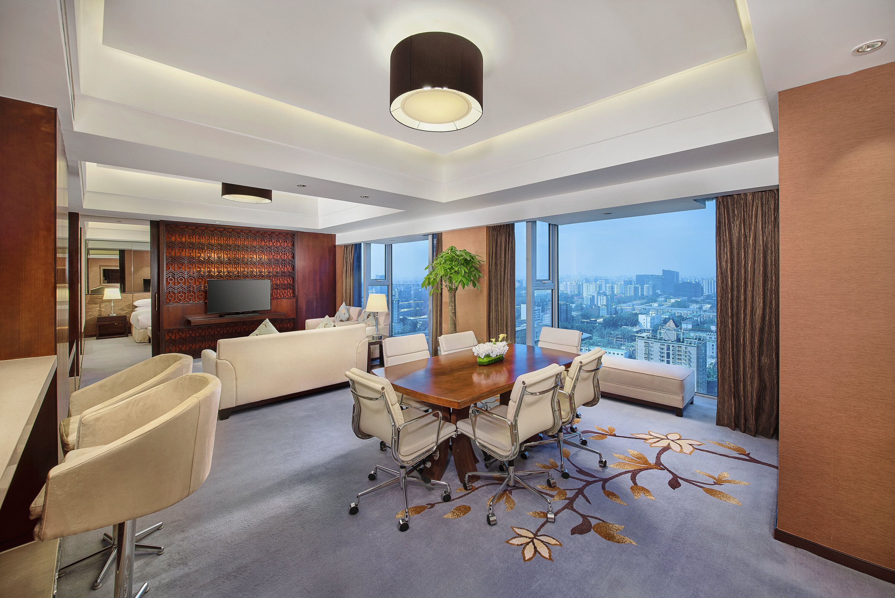 A modern hotel room with a large city view, a dining area with a table and chairs, and a living area with a sofa and armchairs.