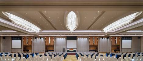 A large conference room with rows of white chairs, a stage with a large screen, and a decorative ceiling design.
