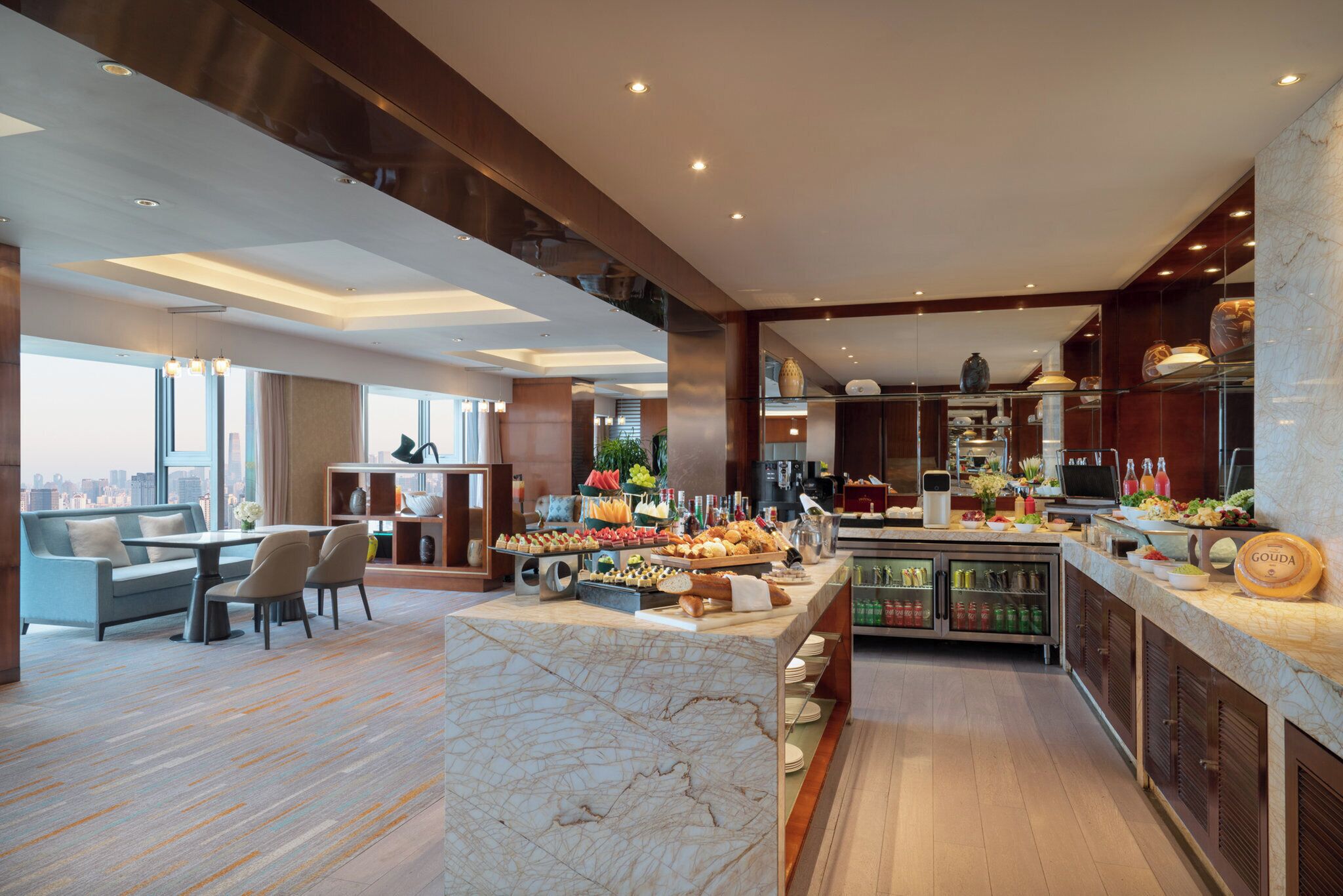 A modern hotel lobby with a seating area, a buffet with various food items, and a view of the cityscape.