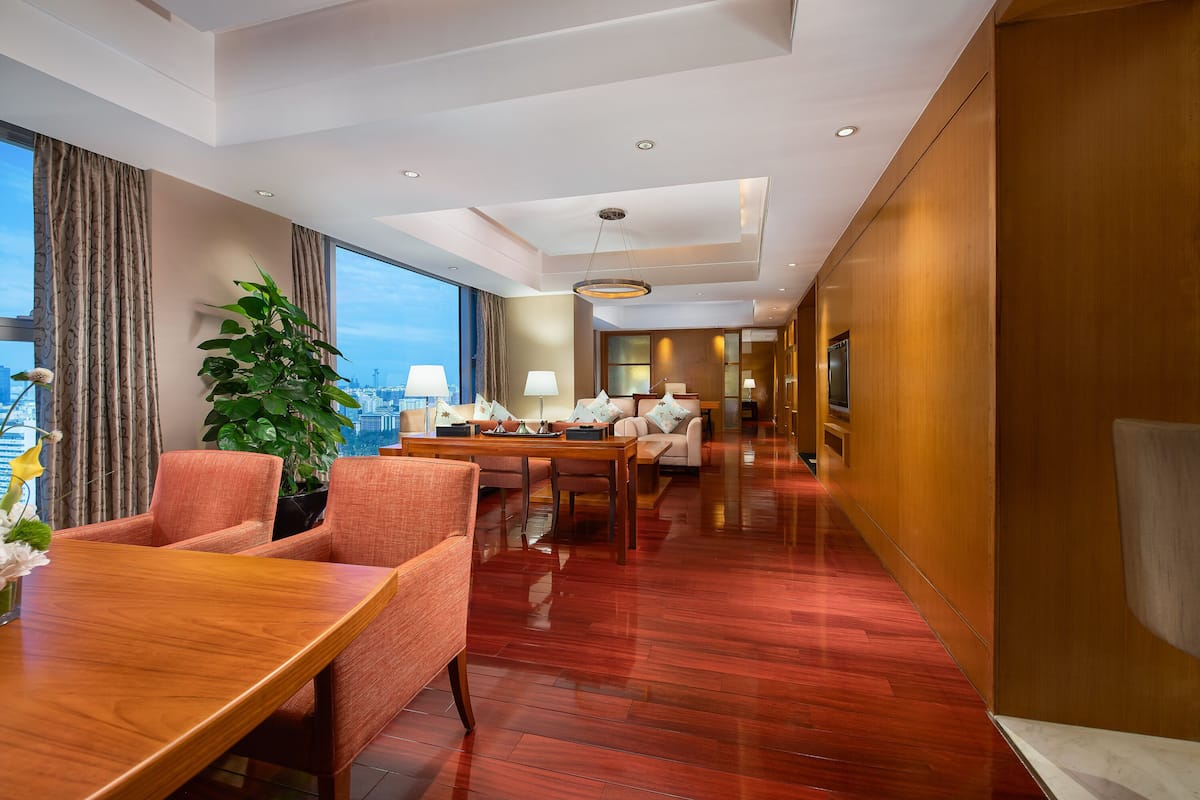 Habitación de hotel con una mesa de comedor de madera, piso cubierto de alfombra roja, una planta grande en maceta y vista a la ciudad a través de la ventana.
