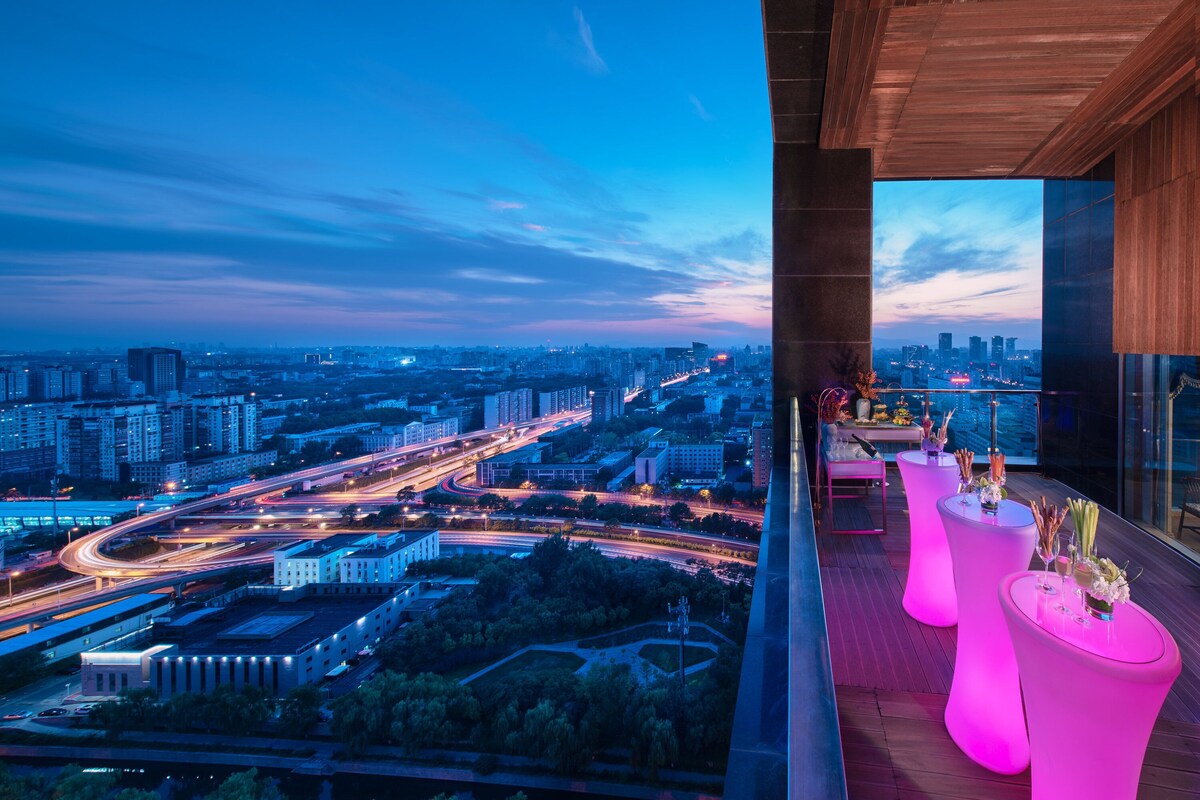A high-rise building with a panoramic city view, a balcony with modern furniture, and a dining area with a table set for two.
