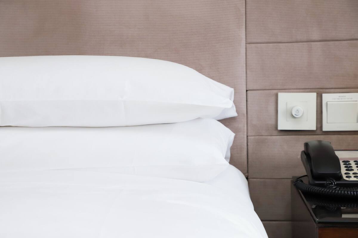 Una cama bien hecha con sábanas blancas, un teléfono negro sobre una mesita de noche de madera, y dos interruptores de luz en la pared.