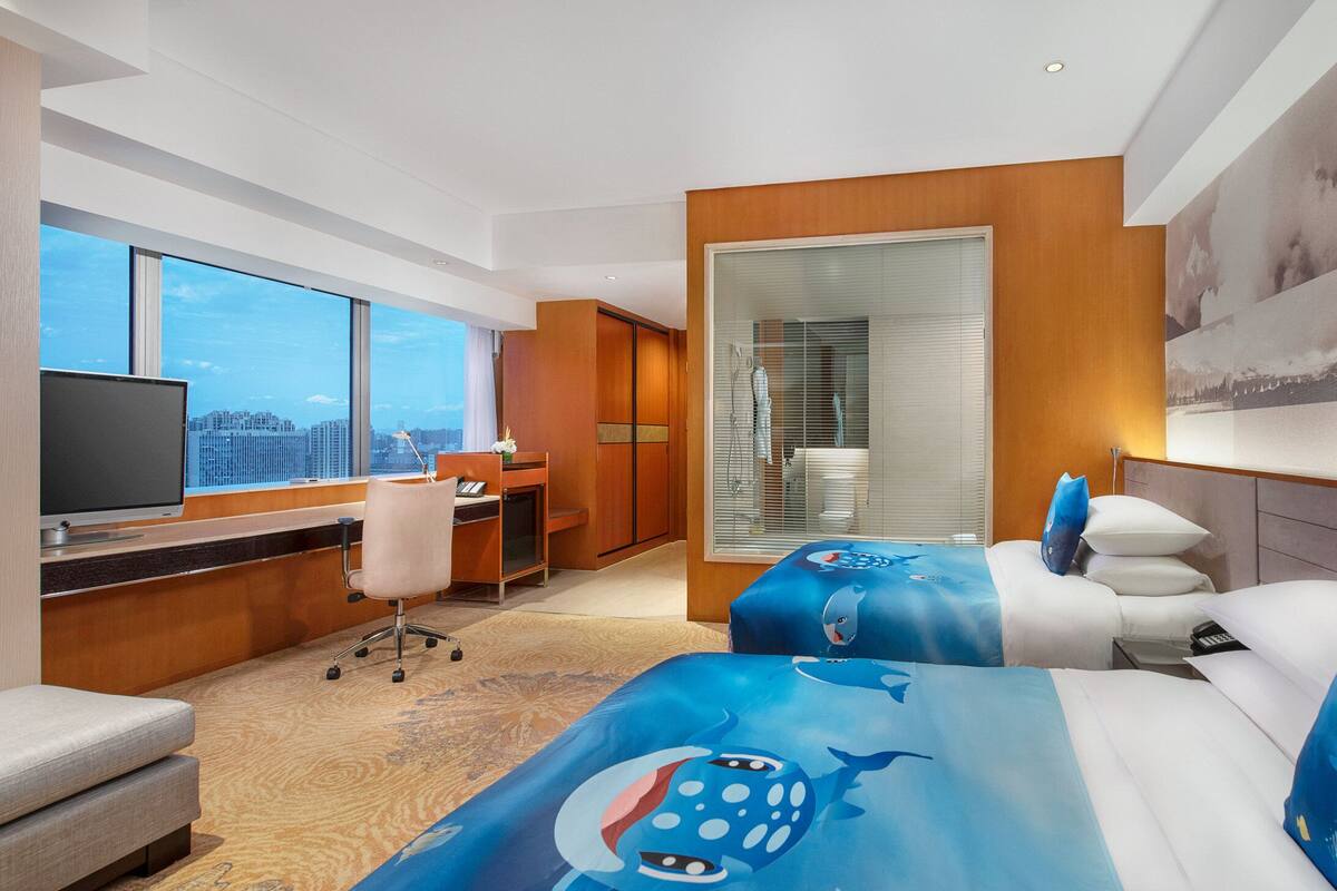 Habitación de hotel con una ventana grande, un escritorio, una silla, una cama con una colcha azul con motivos de tiburón y un televisor.