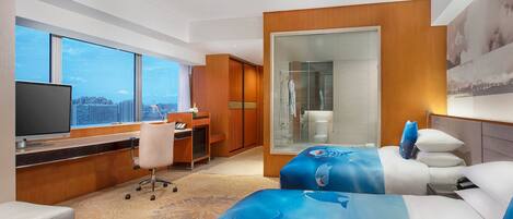 A hotel room with a large window, a desk, a chair, a bed with a blue shark-themed bedspread, and a television.