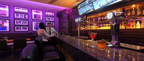 A bar area with a counter, multiple screens displaying sports, framed pictures on the wall, and a variety of bottles and glasses.