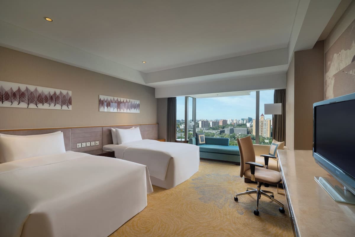 Habitación de hotel con dos camas, un escritorio con silla, un televisor grande y vistas a la ciudad.
