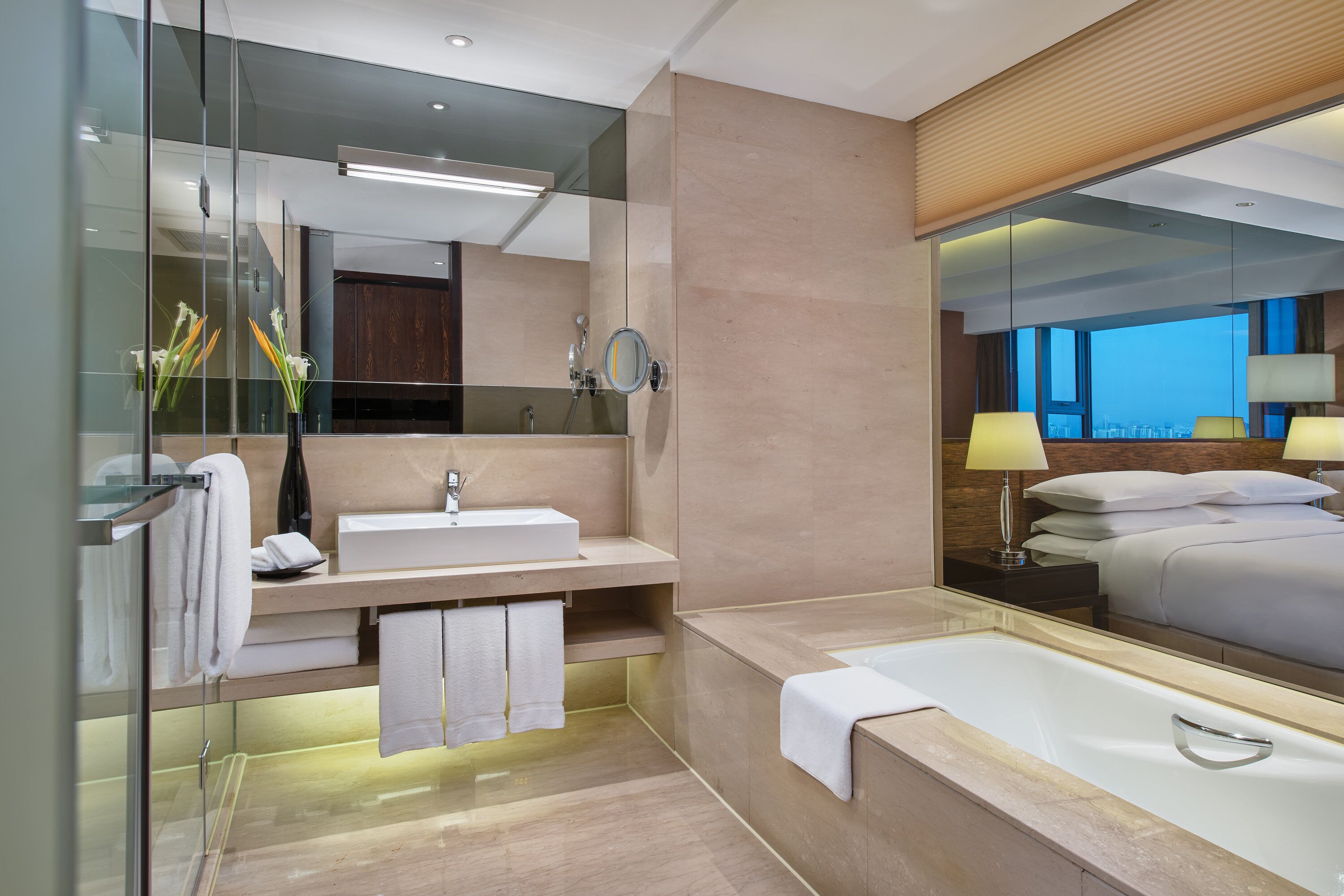 A modern hotel bathroom with a large mirror, a sink, a bathtub, and a view of the cityscape through the window.