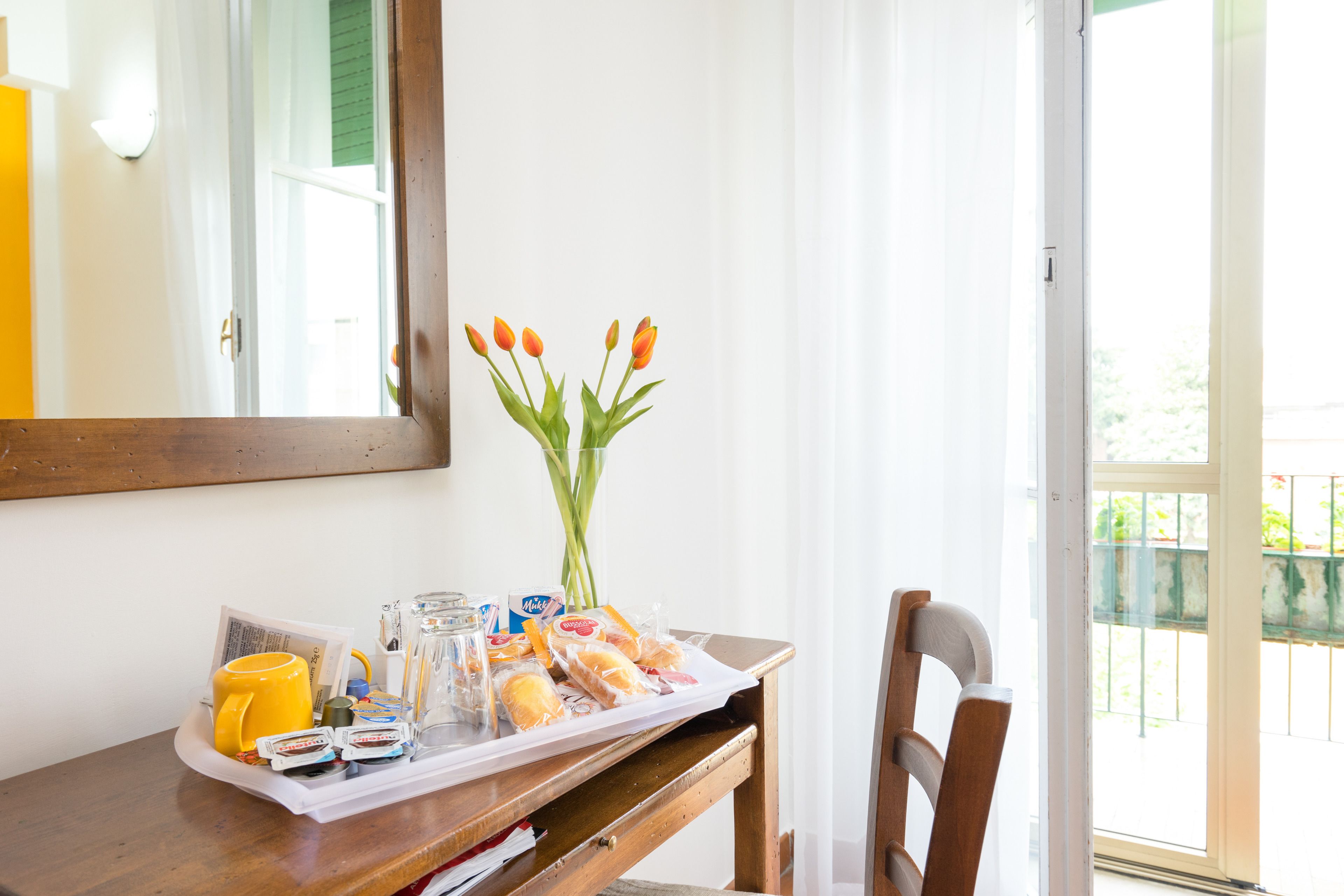 A breakfast tray with food items, a vase with flowers, and a yellow mug on a wooden table near a window.