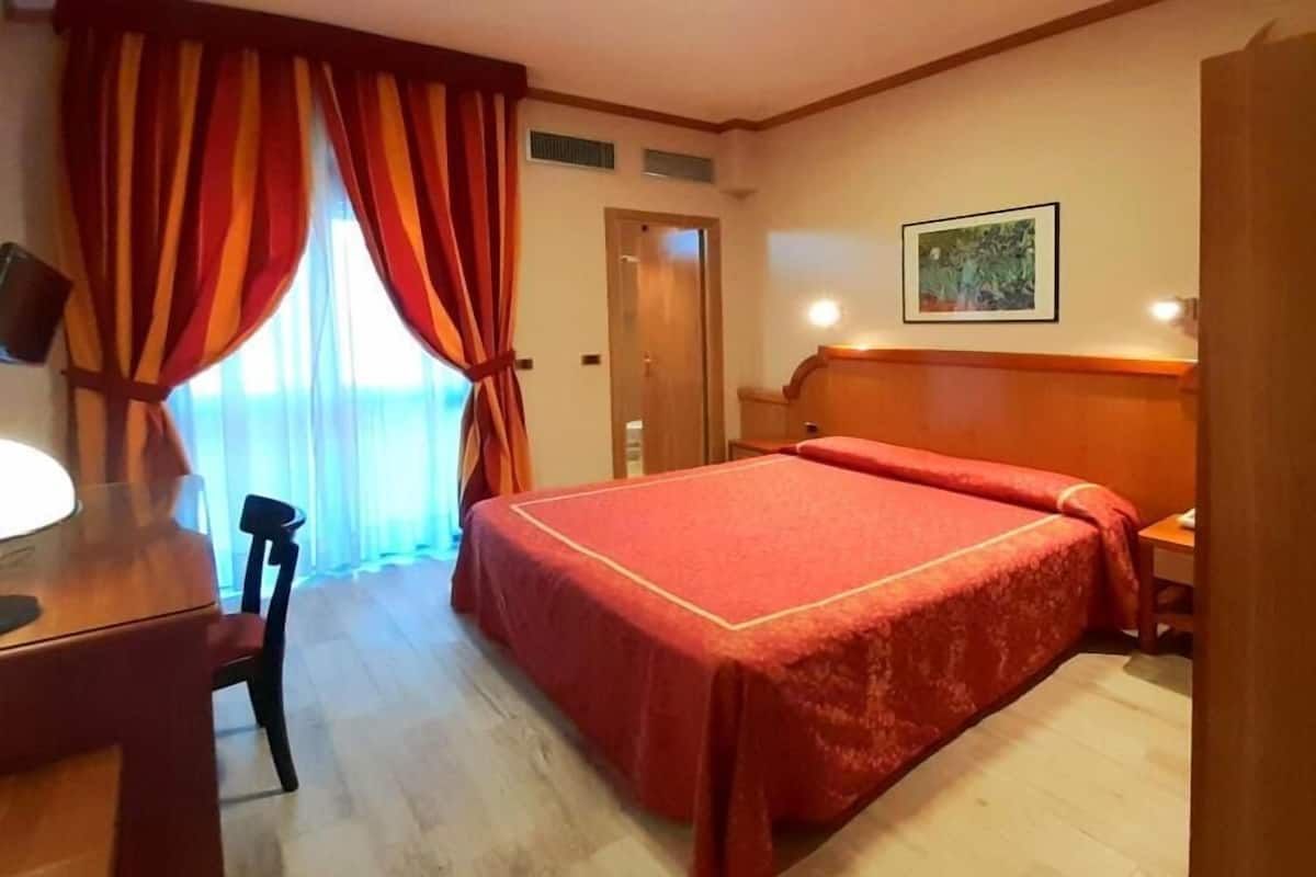 A hotel room with a bed, a desk, a chair, a nightstand, and a door.