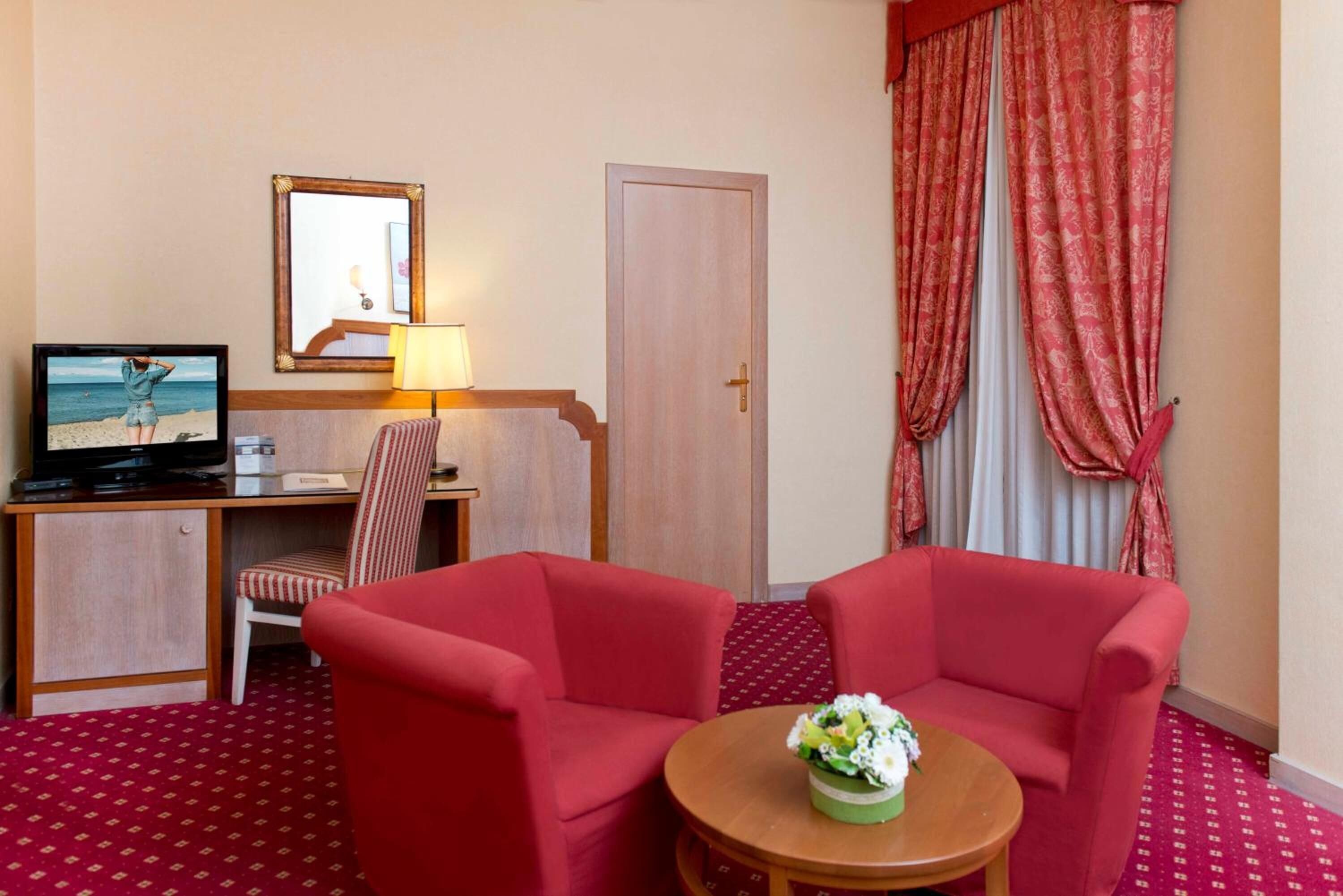 A hotel room with a desk, a television, a chair, two red armchairs, a small round table with a flower vase, and red patterned carpet.