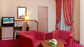 A hotel room with a desk, a television, a chair, two red armchairs, a small round table with a flower vase, and red patterned carpet.