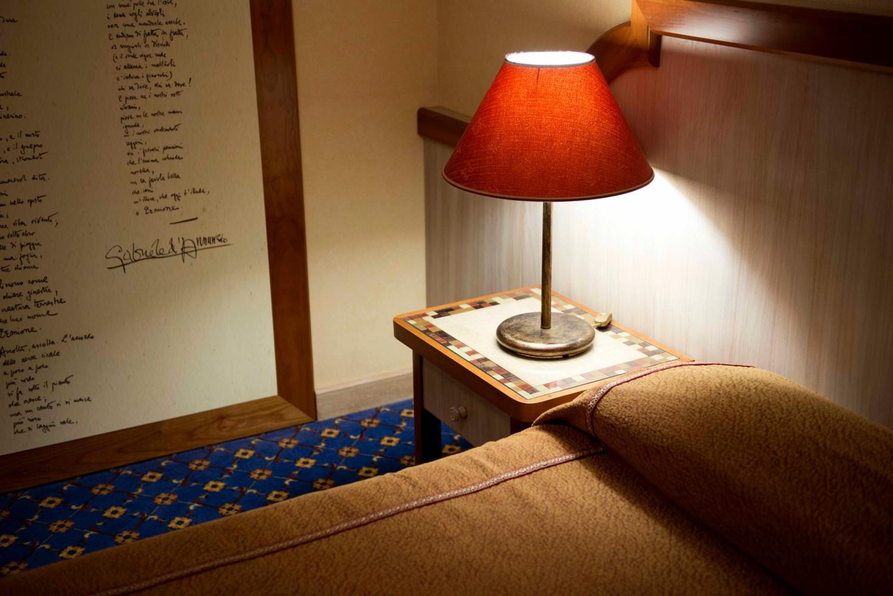 A room with a small table, a lamp, a blue patterned rug, and a wall with a list of names.
