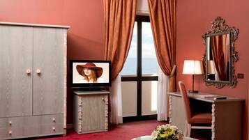 Habitación de hotel con un armario grande, televisión, escritorio con lámpara, silla y vistas al mar a través de una ventana con cortinas.