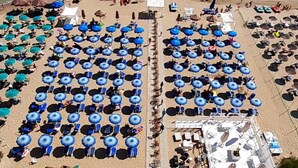 En strand med rader av blå parasoller och stolar, ett poolområde och en stenmur som vågbrytare i bakgrunden.