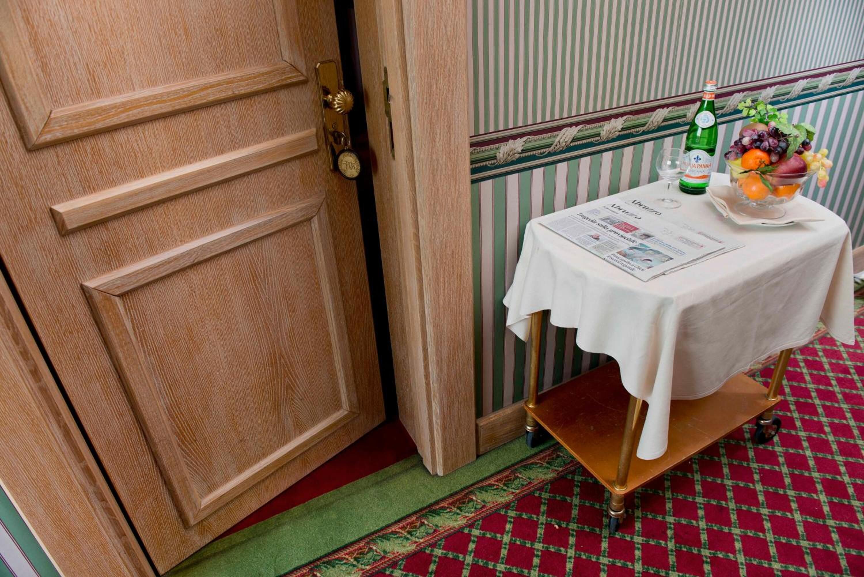 A wooden door with a brass handle, a small table with a white tablecloth, a bottle of water, a glass, and a bowl of fruit.