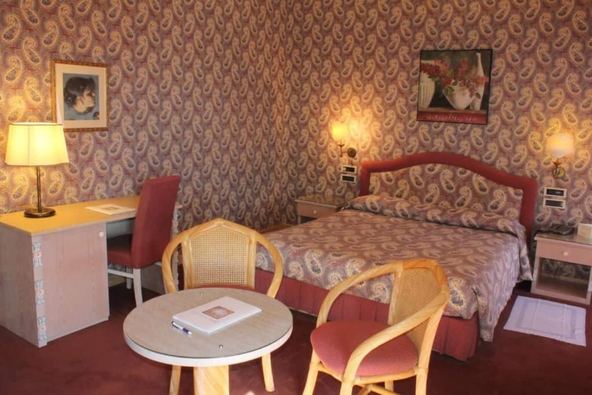 A hotel room with a patterned wallpaper, a bed with a headboard, two bedside tables, a desk with a chair, and a round table with two chairs.