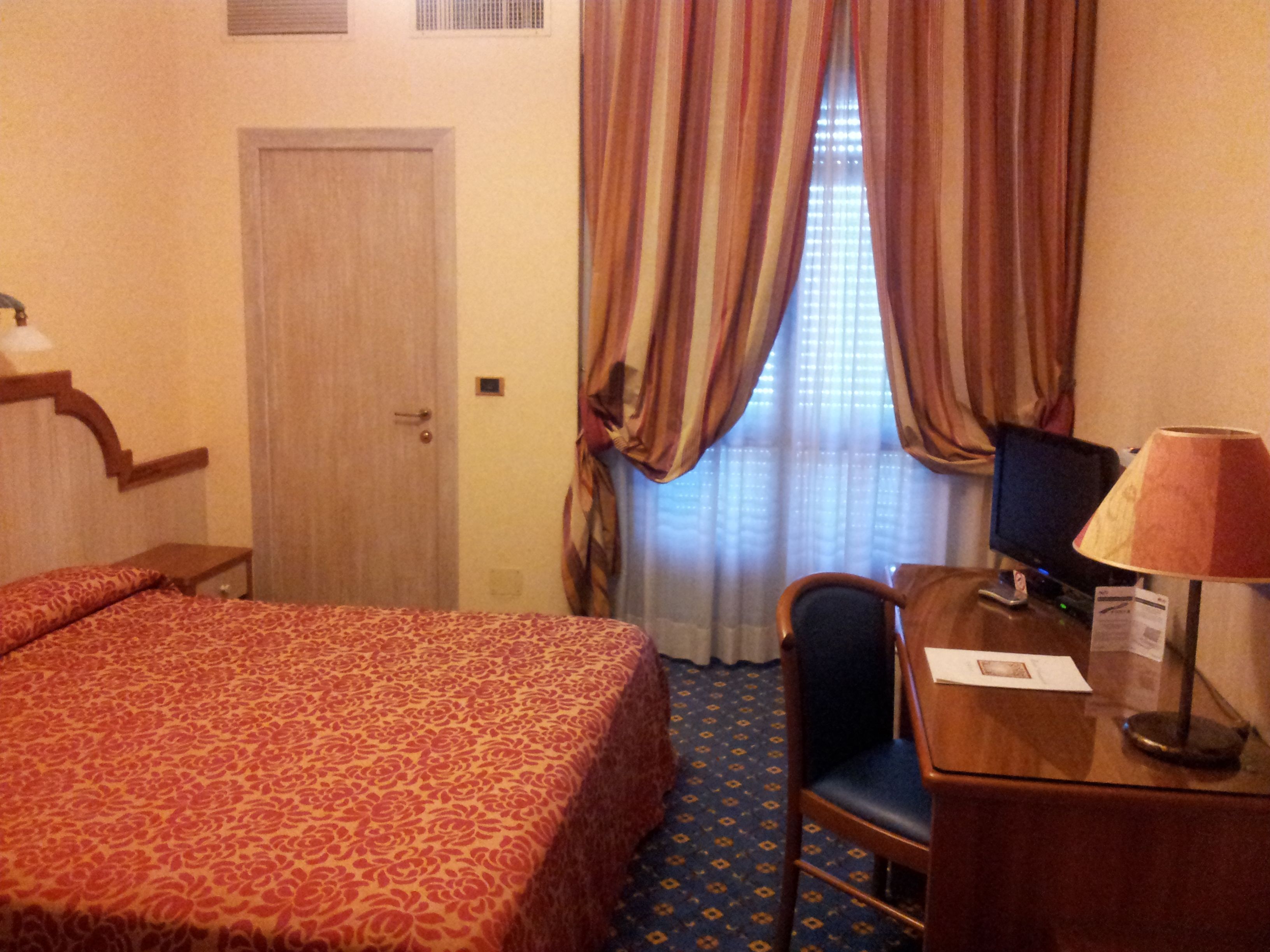 A hotel room with a bed, a desk, a chair, a lamp, and a television.