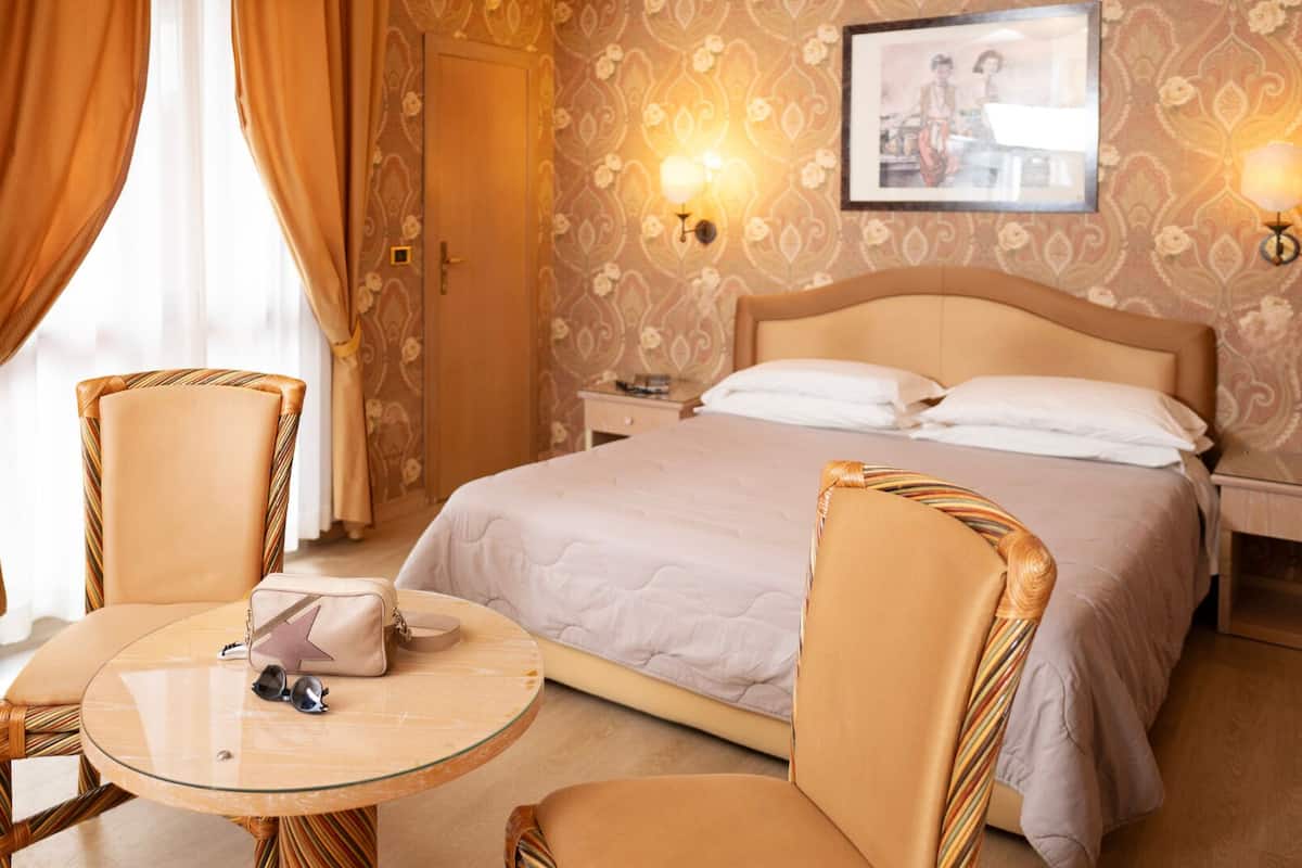 A hotel room with a bed, two nightstands, a bedside table with a lamp, a chair, and a window with curtains.