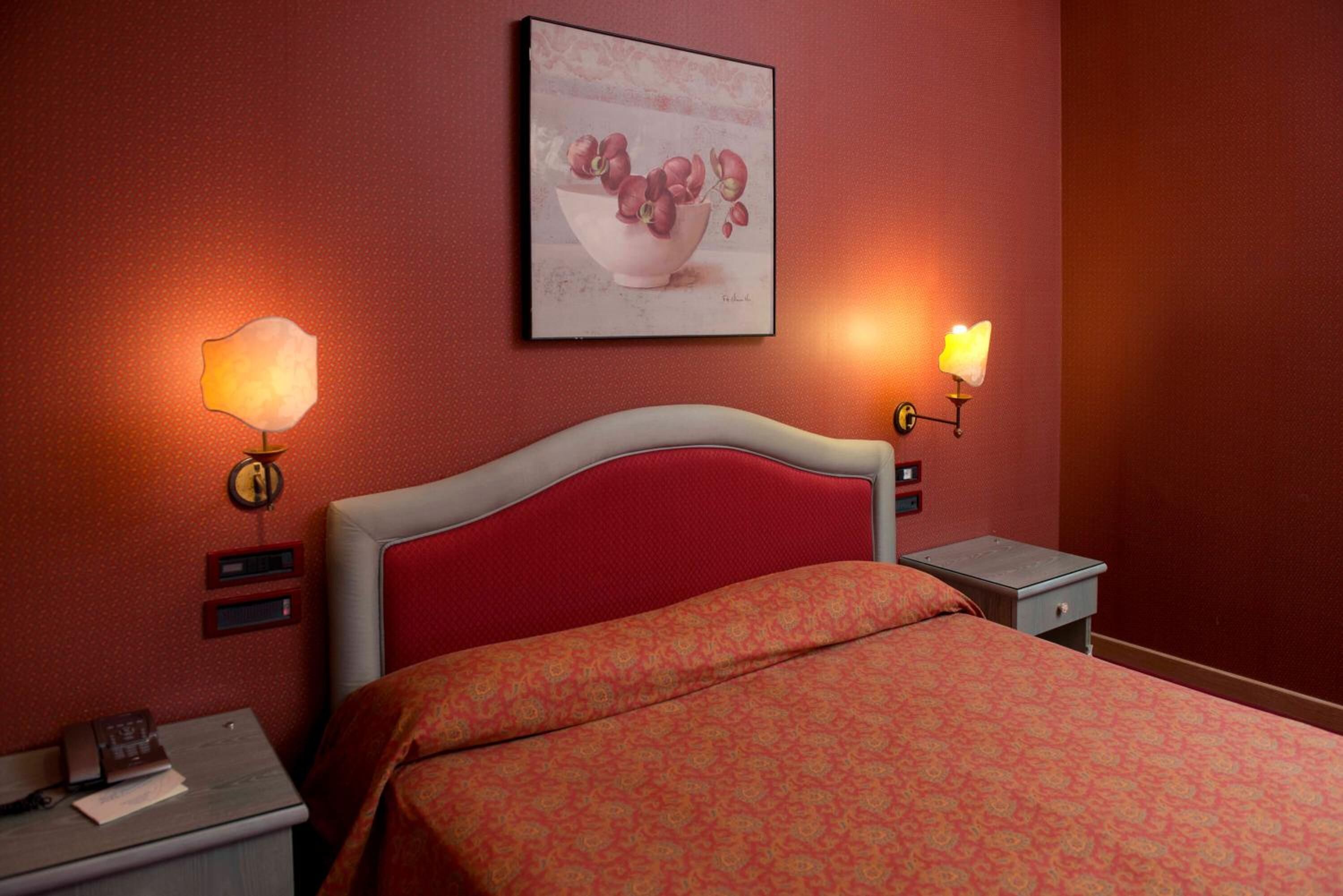 A hotel room with a red headboard, a bed with a patterned bedspread, two bedside tables with lamps, and a framed picture on the wall.
