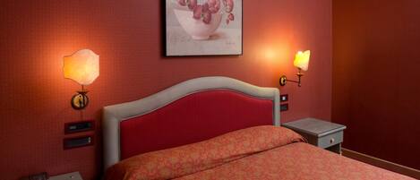 A hotel room with a red headboard, a bed with a patterned bedspread, two bedside tables with lamps, and a framed picture on the wall.