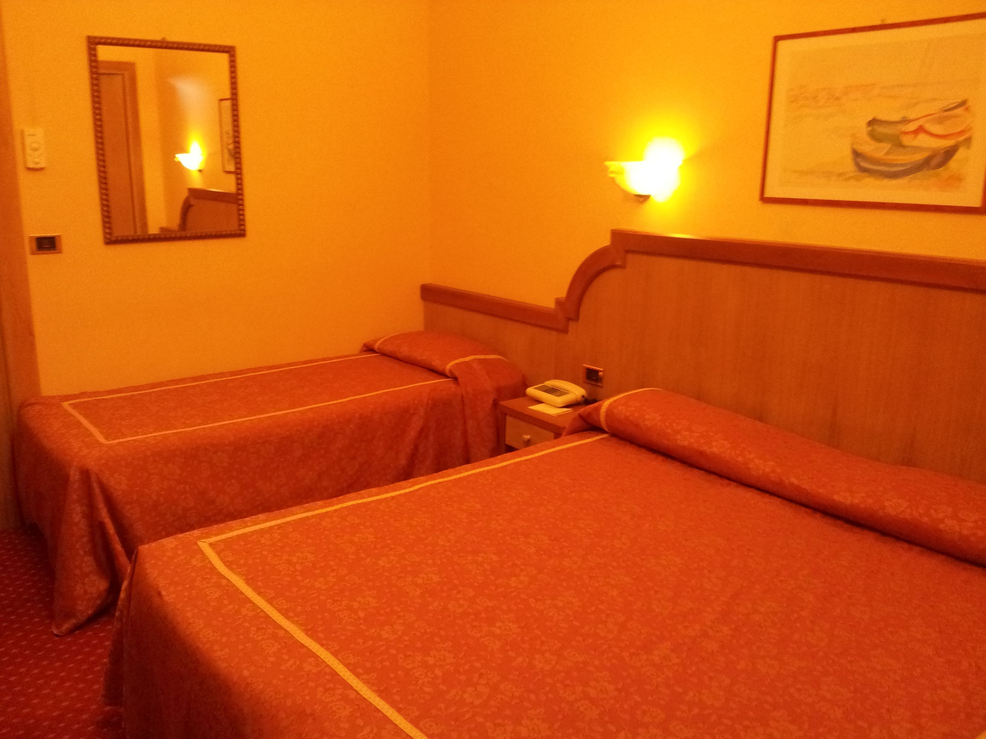 Hotellihuone, jossa on kaksi erillistä sänkyä, yöpöytä lampun kanssa, puhelin ja seinällä oleva kehystetty kuva.
