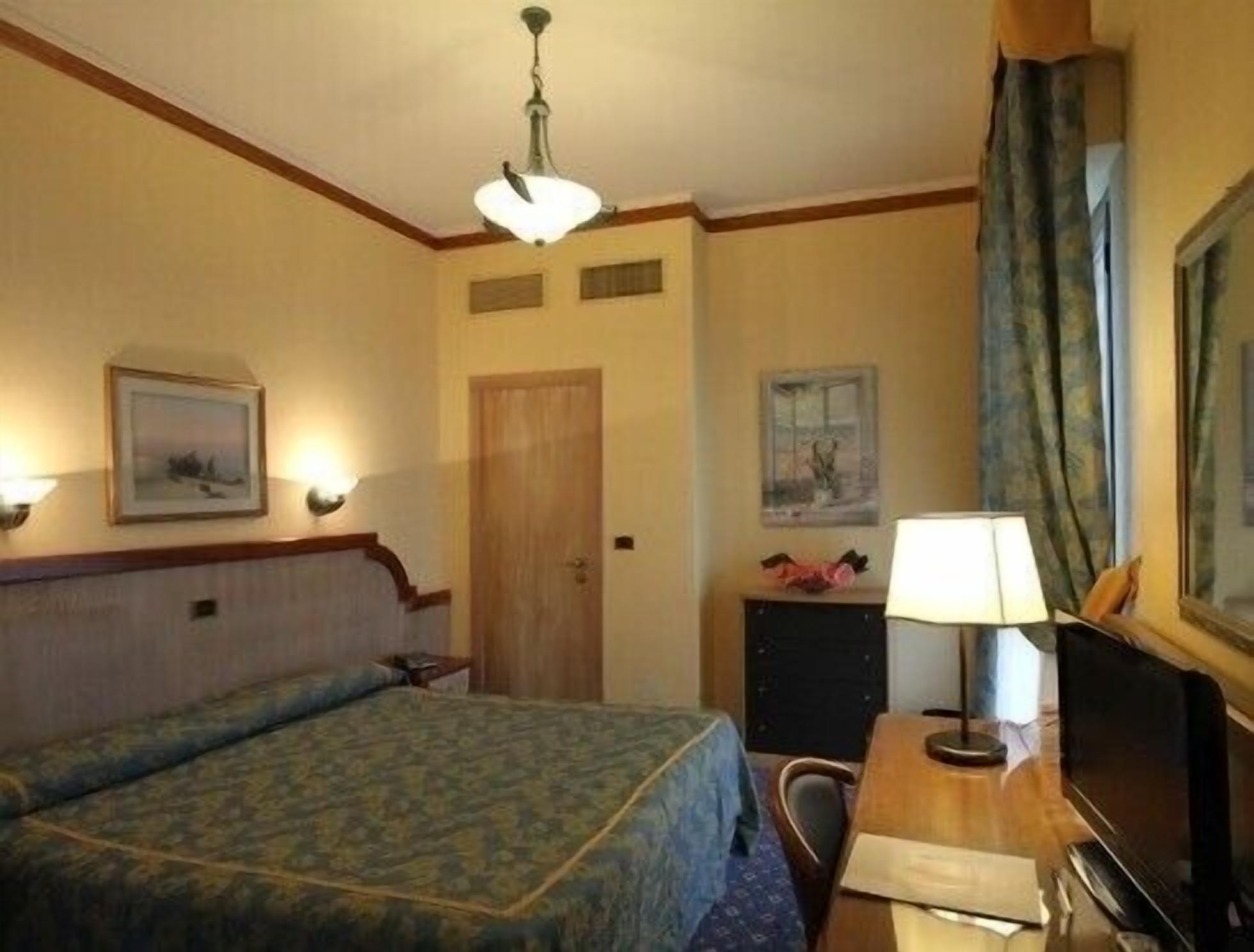 A hotel room with a bed, a desk, a chair, a lamp, and two framed pictures on the wall.