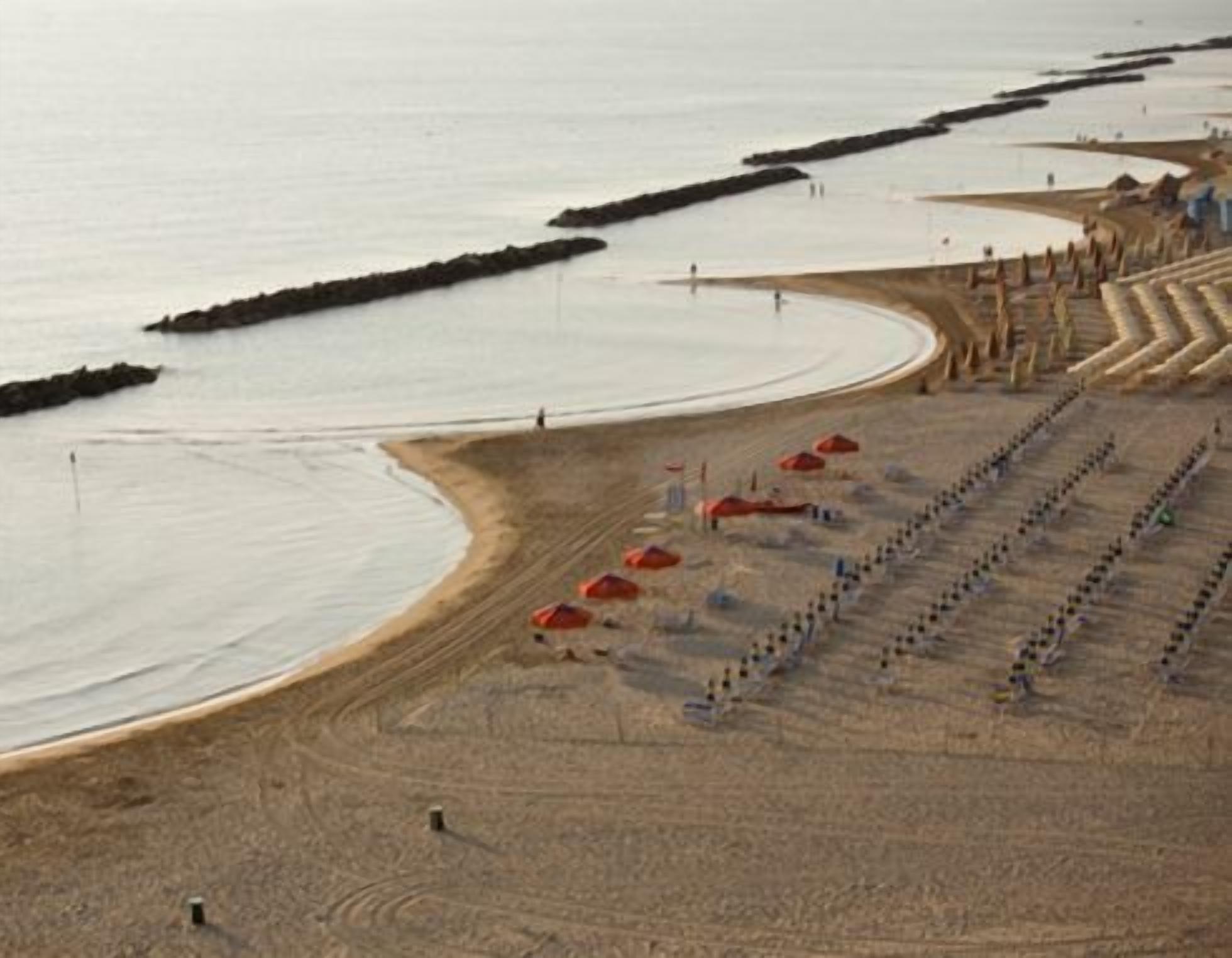 Een strand met een lange rij parasols en ligbedden, een gebogen kustlijn en een steiger die het water in loopt.