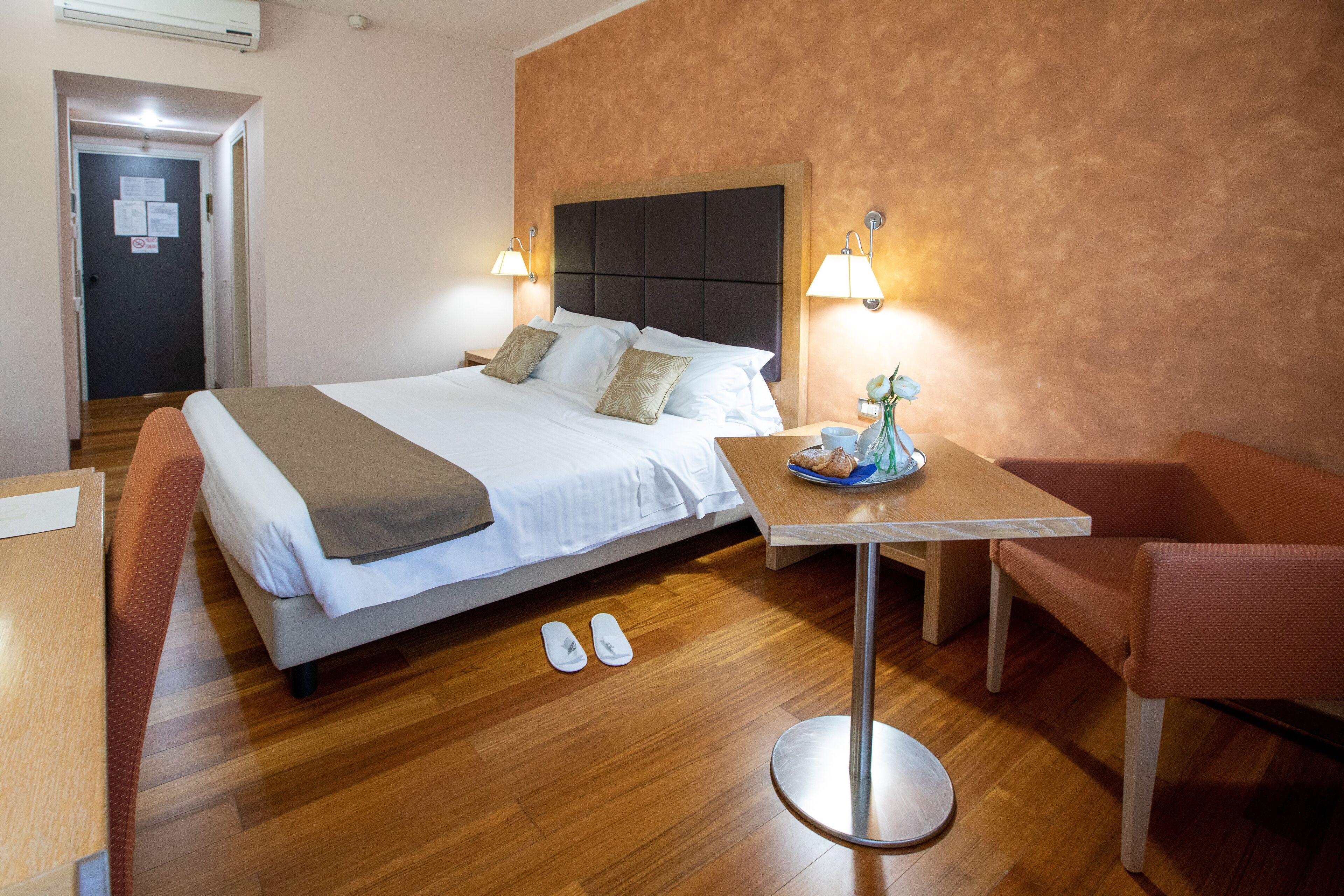 A hotel room with a bed, a table, and a chair. There are slippers on the floor.