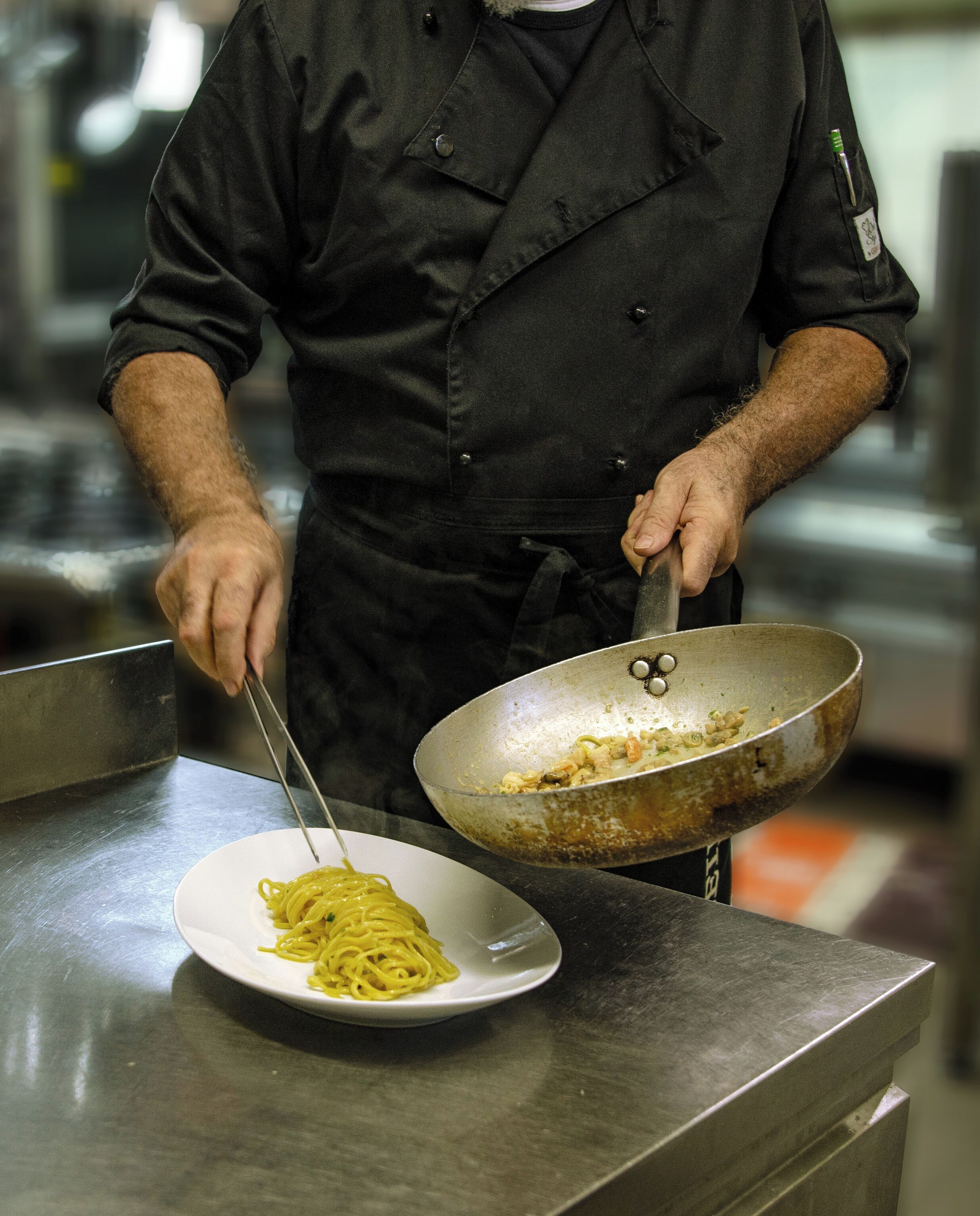 En kok i sort uniform holder en wok med mad og står ved siden af en tallerken med spaghetti.