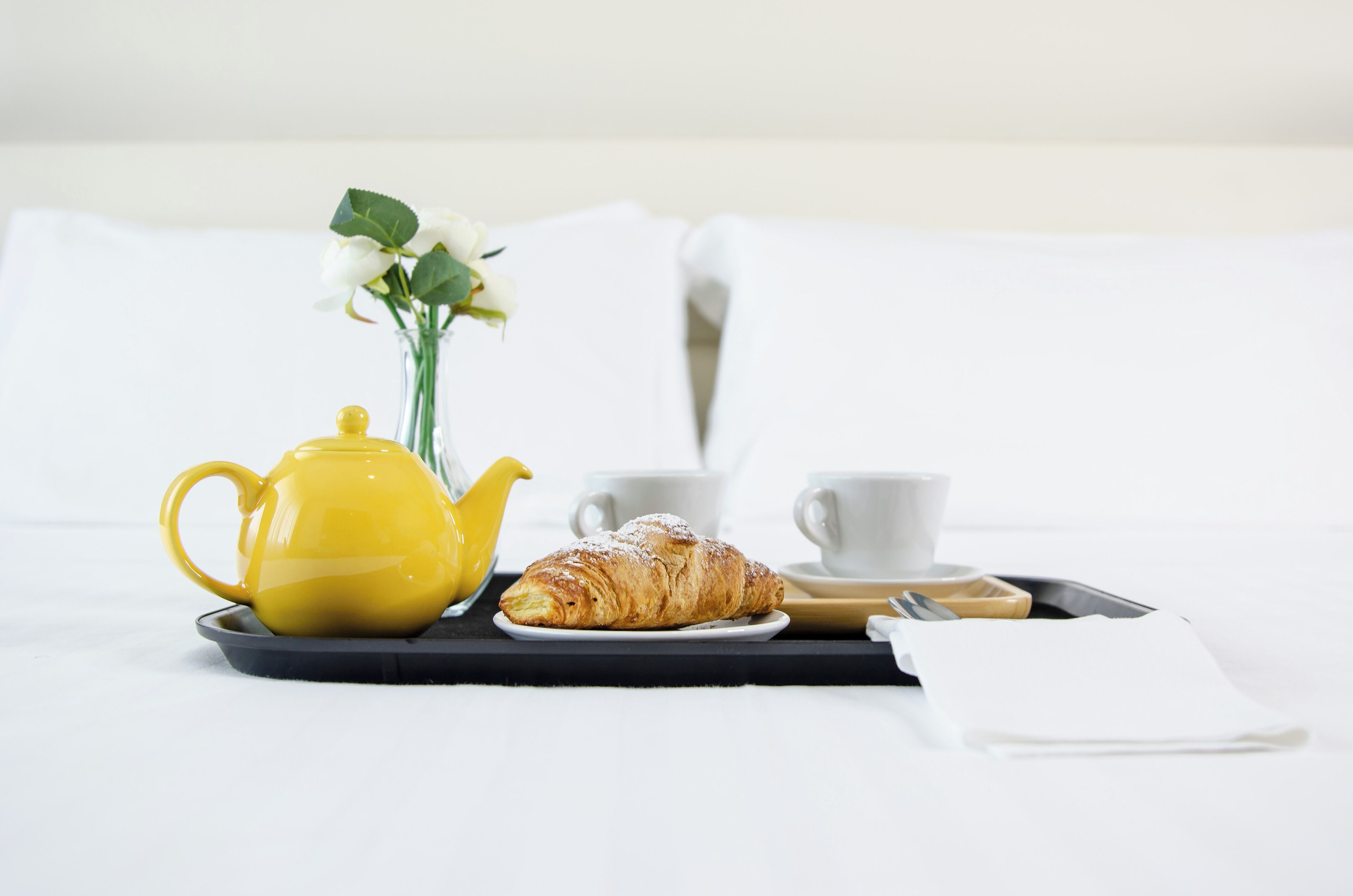 Una bandeja de desayuno con una tetera amarilla, un croissant y una taza blanca sobre una cama con ropa de cama blanca.