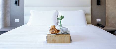A neatly made bed with white linens, a wooden block with a cup and pastries, and a vase with a flower.