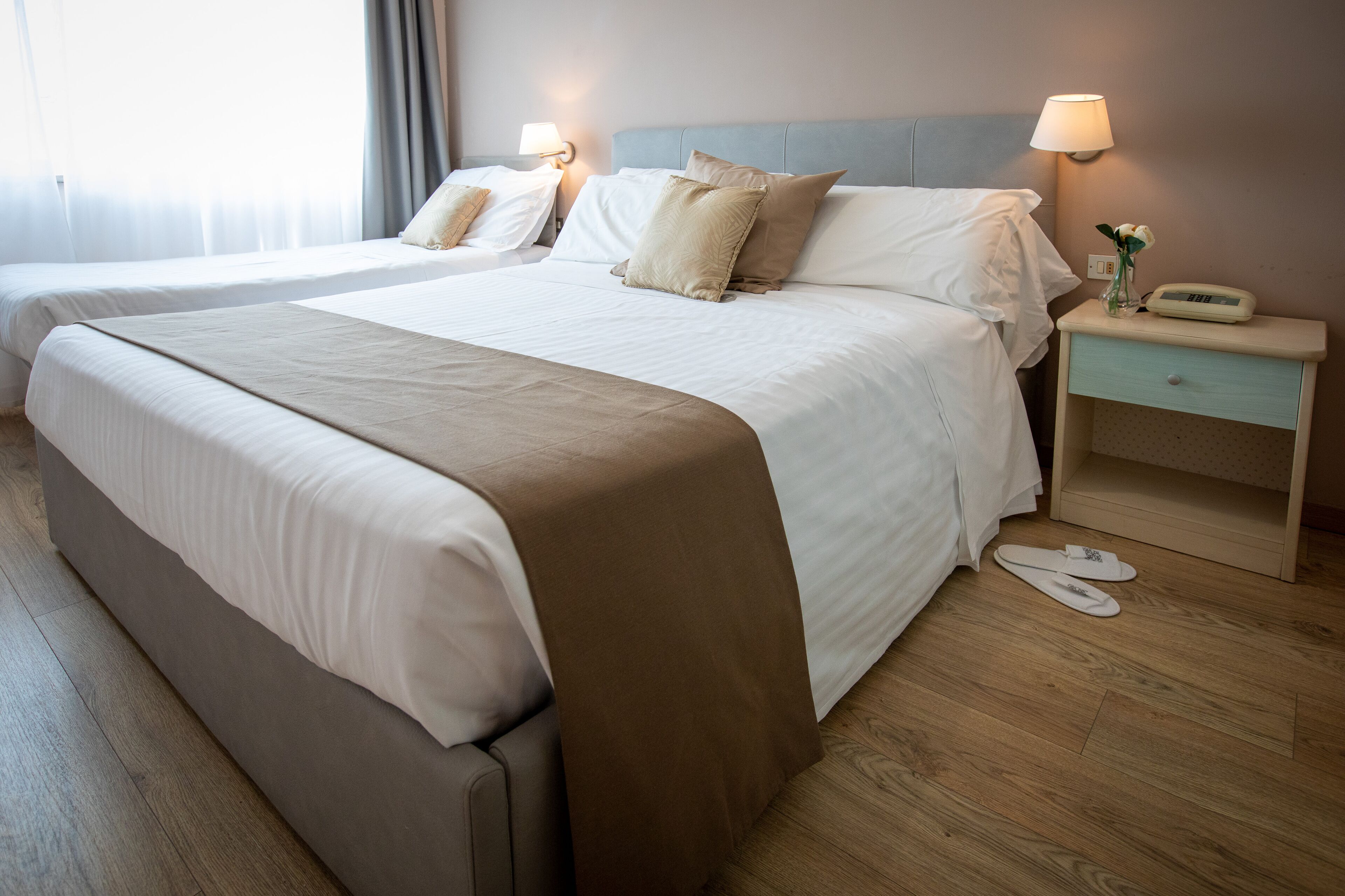 A hotel room with two beds, a bedside table with a lamp, a clock, and a pair of slippers on the floor.