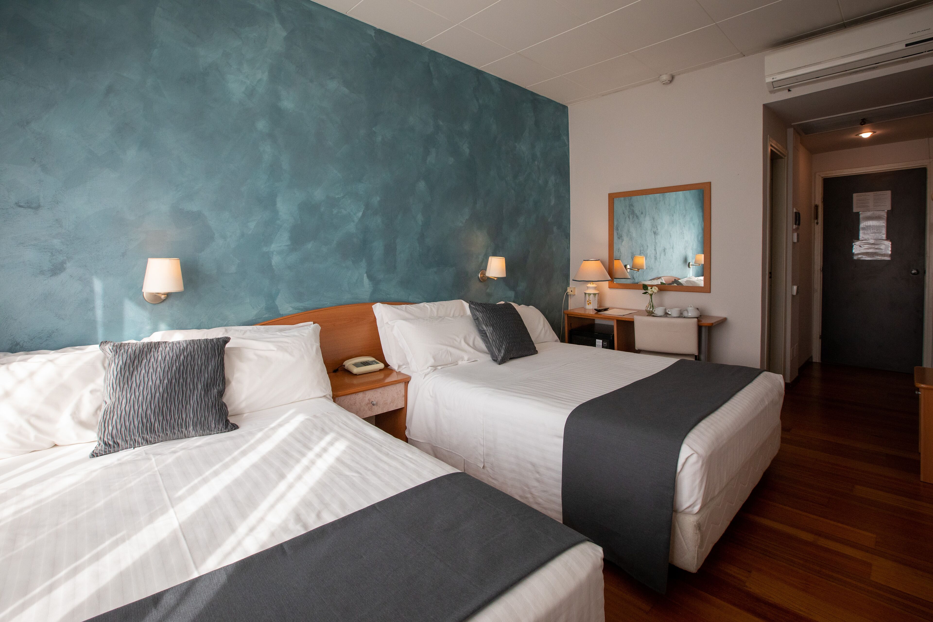 A hotel room with two beds, a teal accent wall, bedside lamps, a nightstand, a mirror, and a wooden floor.