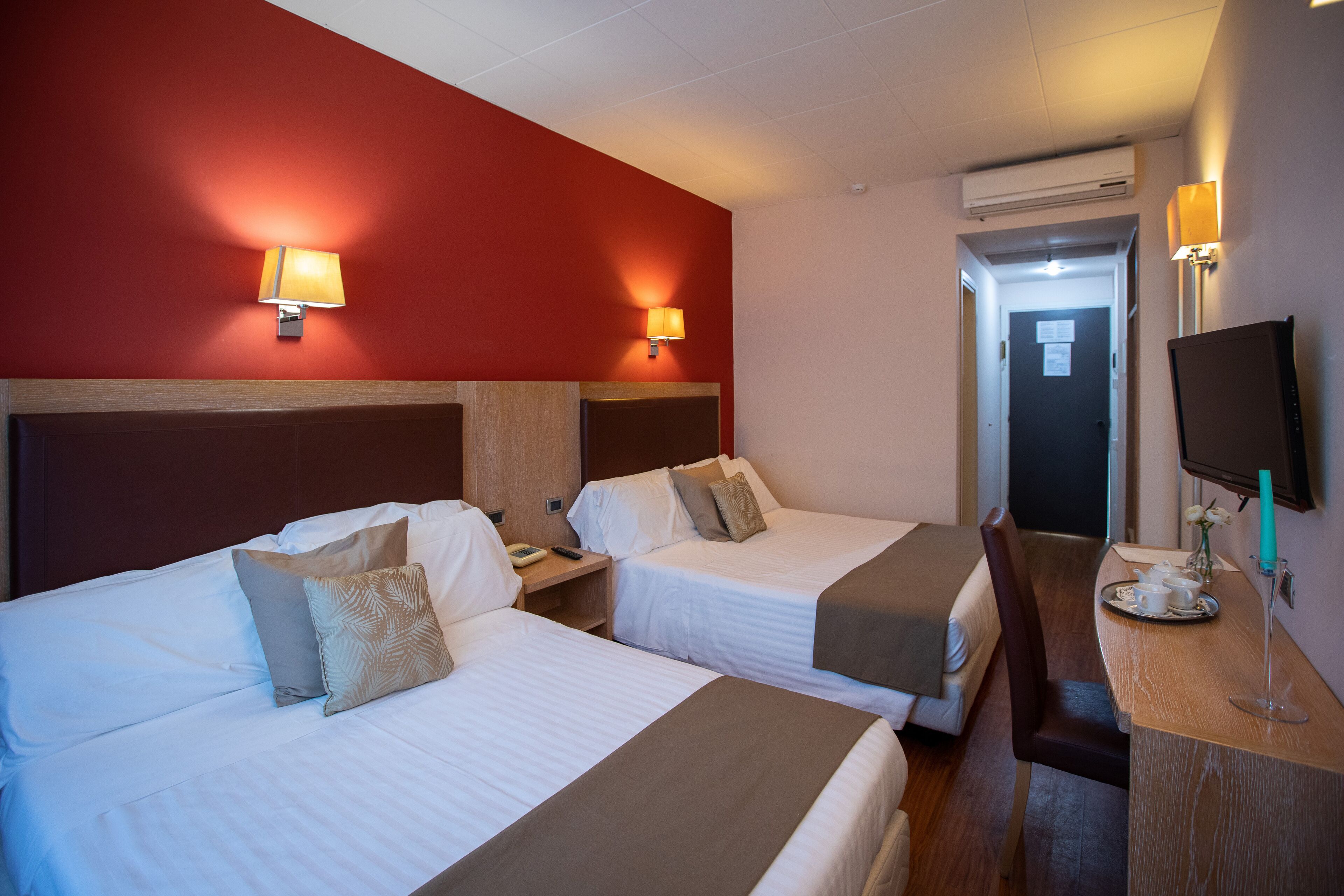 Habitación de hotel con dos camas, un escritorio con silla, un televisor y una puerta que da a otra habitación.
