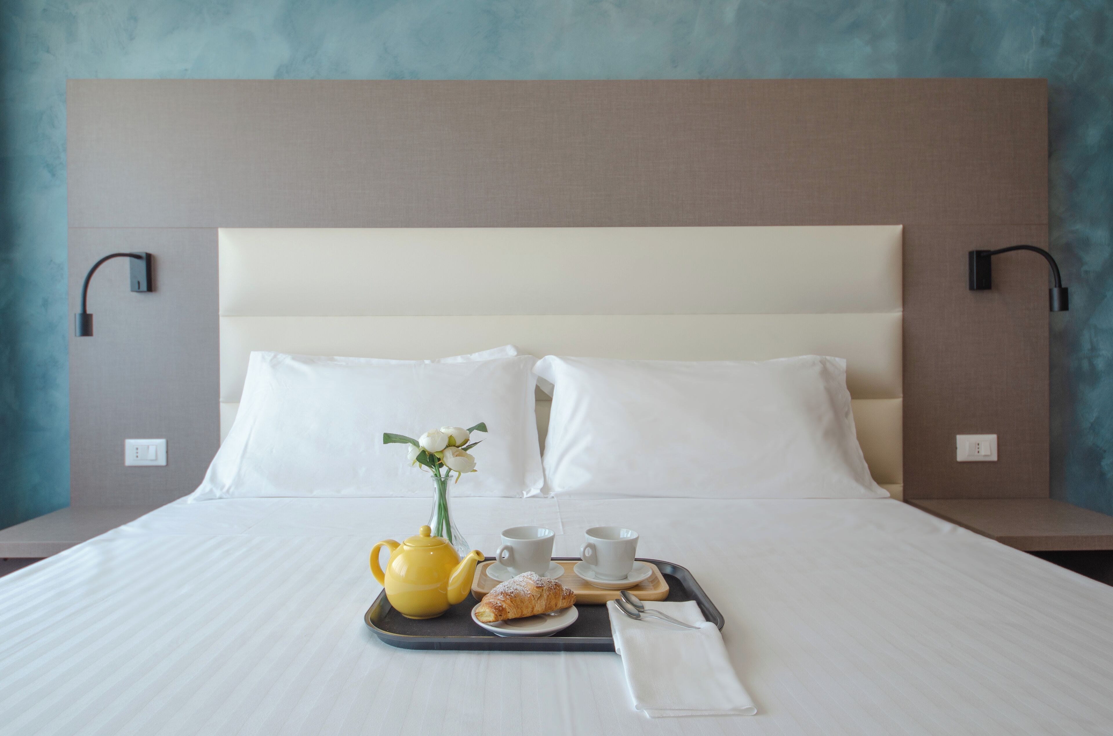 A neatly made bed with white linens, a tray with a teapot, cups, a pastry, and a napkin.