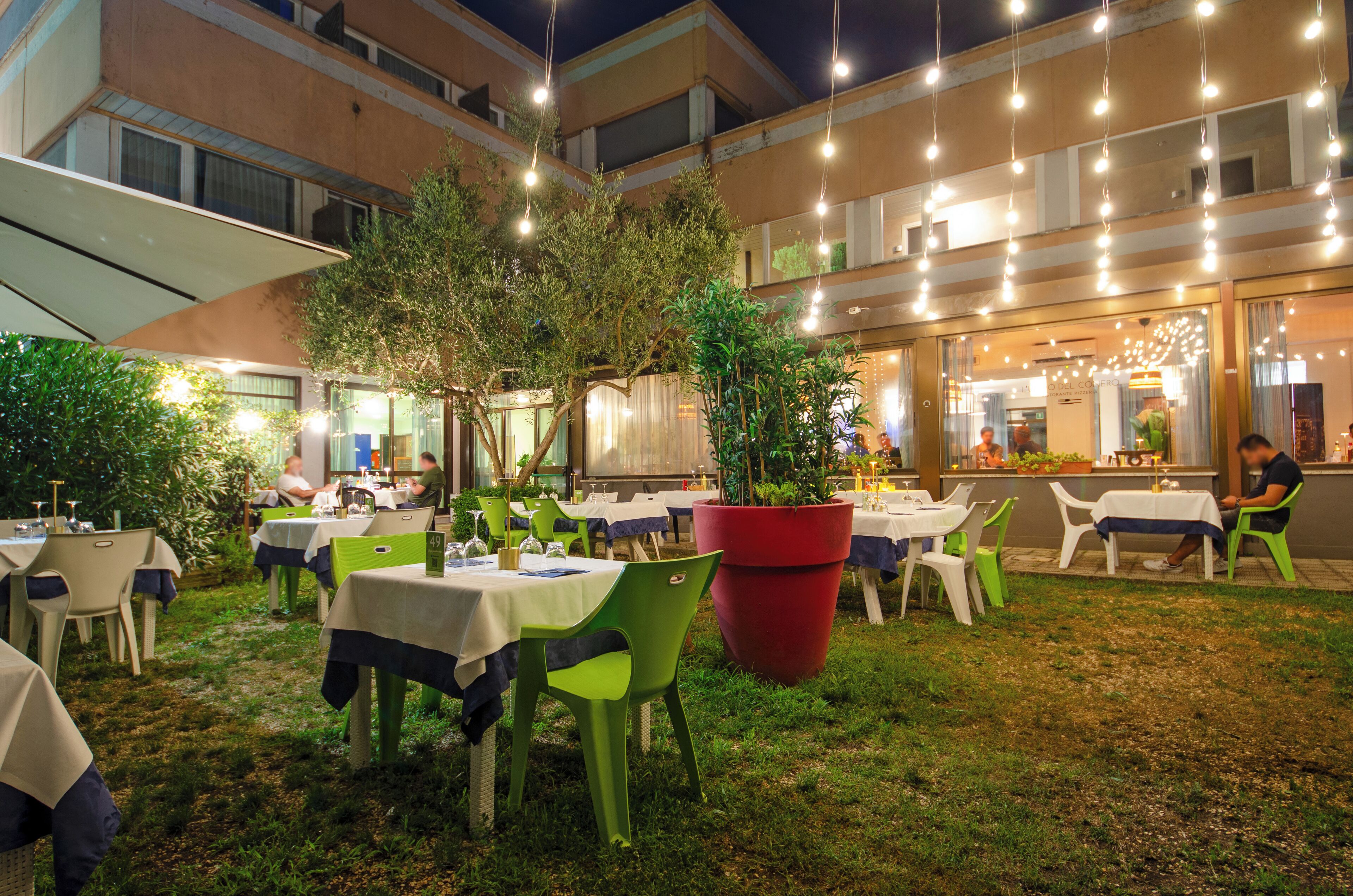 Un espacio de comedor al aire libre con mesas y sillas, rodeado de follaje y un edificio al fondo.