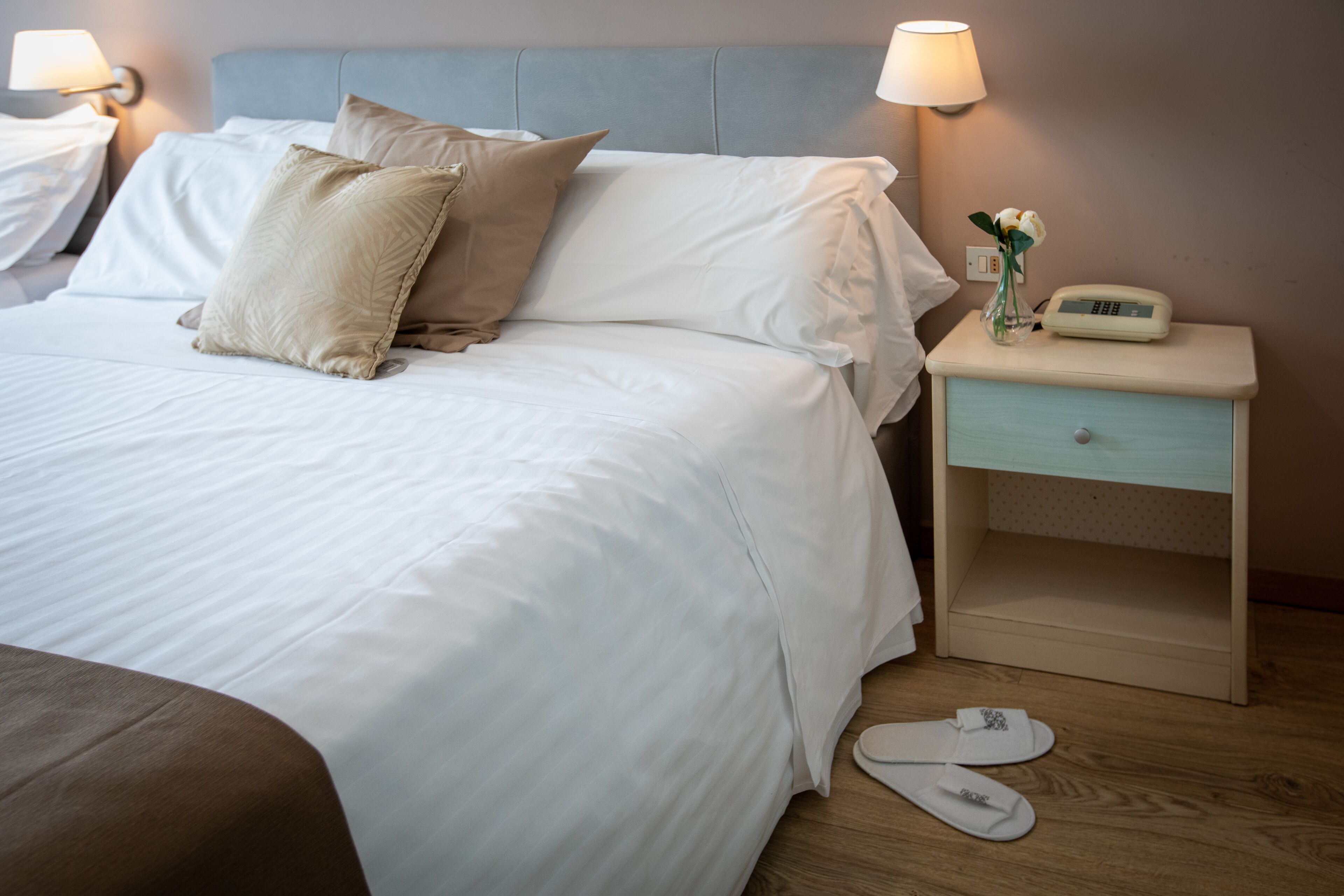 A neatly made bed with white linens and beige pillows, a bedside table with a lamp, a small vase with flowers, and a bedside scale.