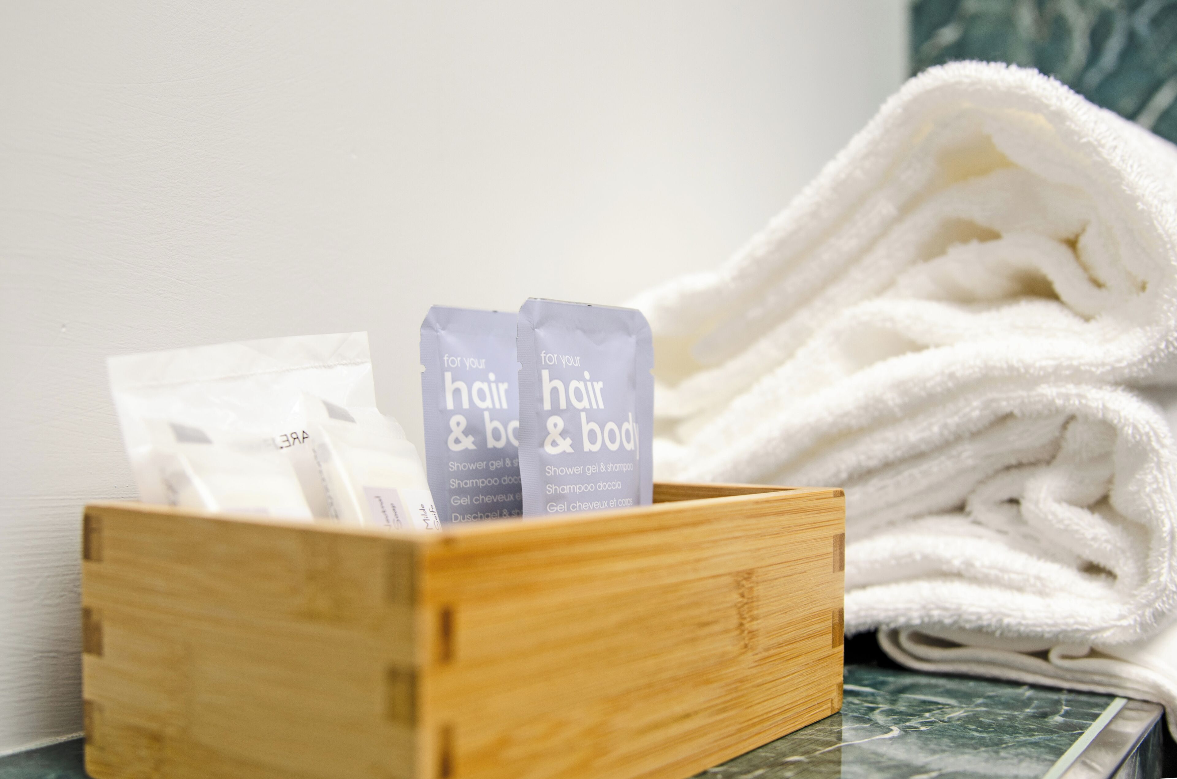 A wooden box containing two toiletry bags labeled "for your hair & body" and a stack of white towels.